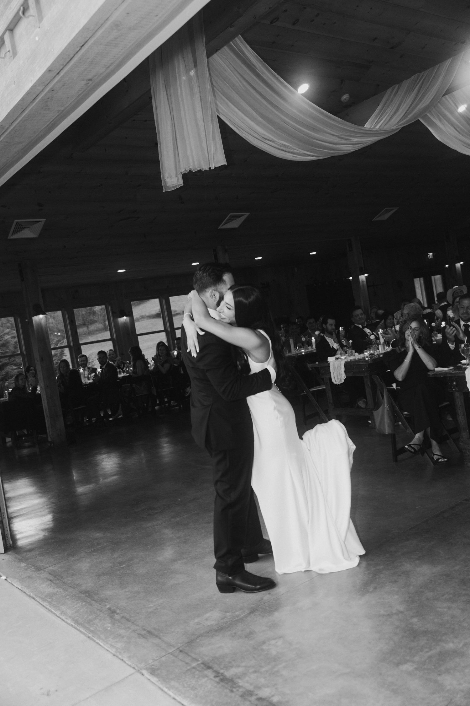 Bride and groom share their first dance in a decorated wedding reception hall while guests applaud.