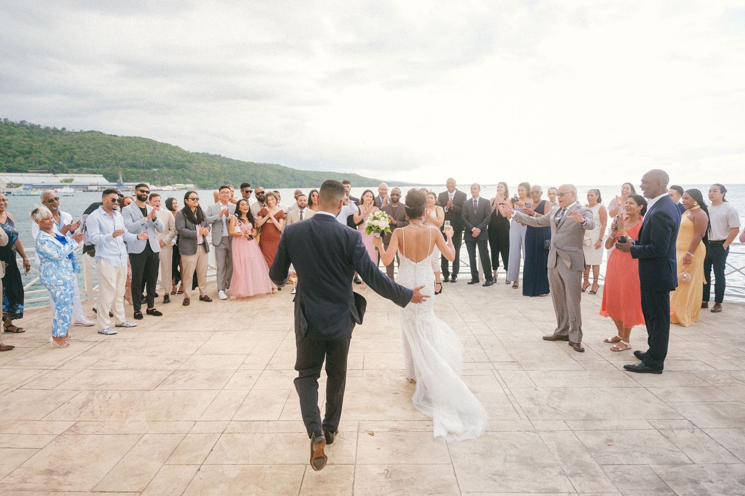Wedding guests form a circle around a couple dancing on an outdoor terrace overlooking water.