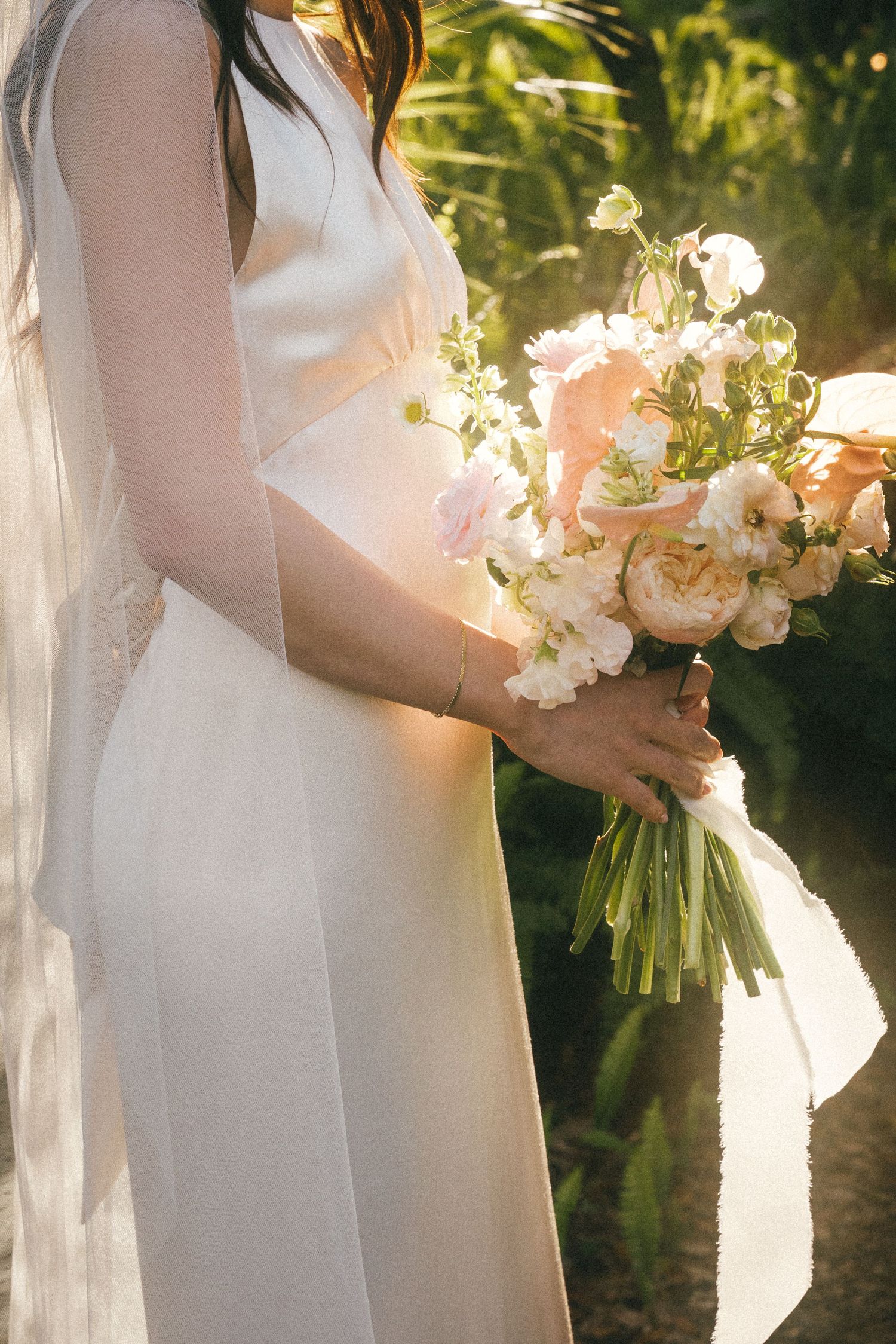 Elegant white bridal bouquet held against a flowing wedding dress in natural sunlight.