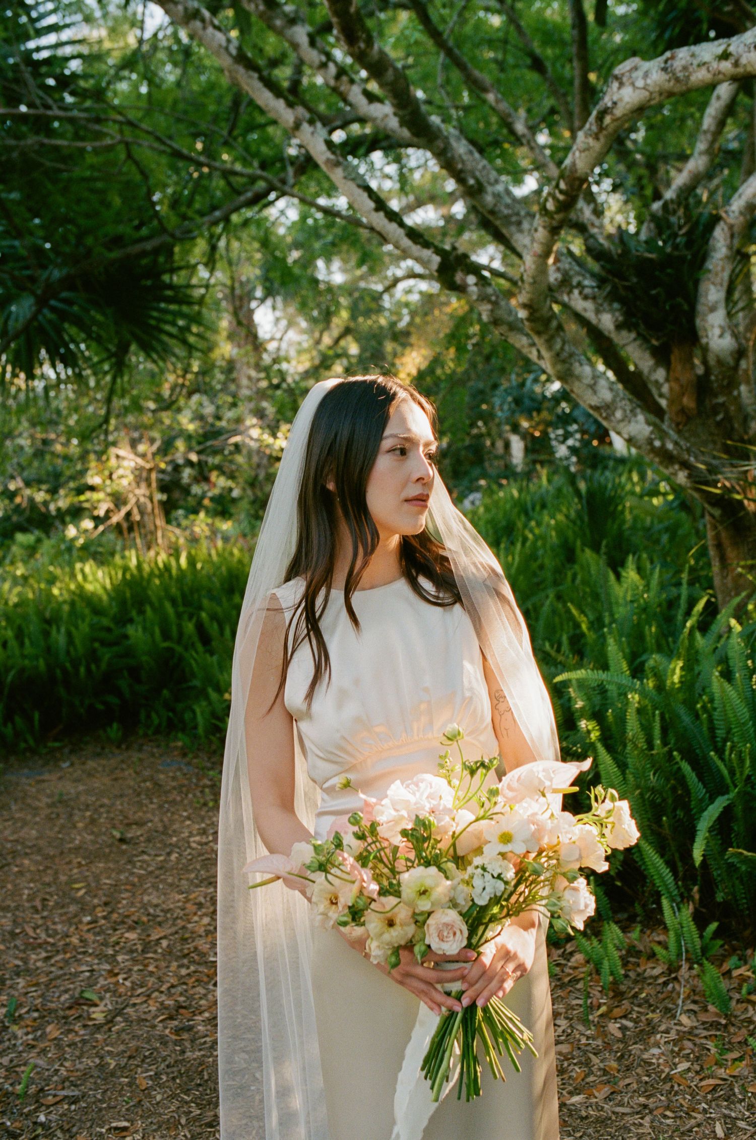 A bride in a flowing cream dress holds a pastel bouquet in a garden setting.