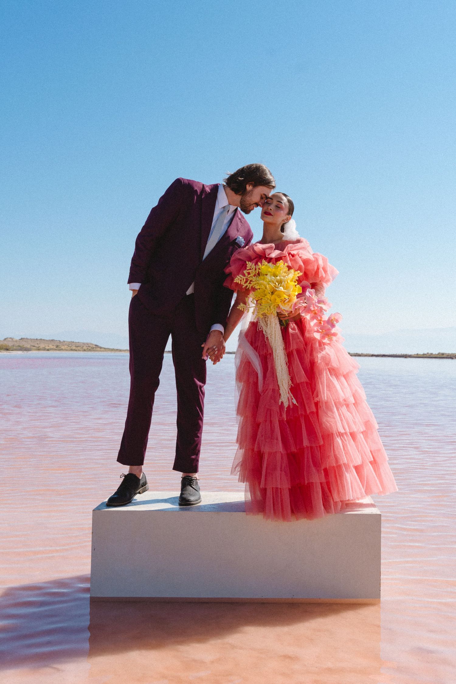 Wedding couple in formal attire posing on white platform in pink salt lake landscape.