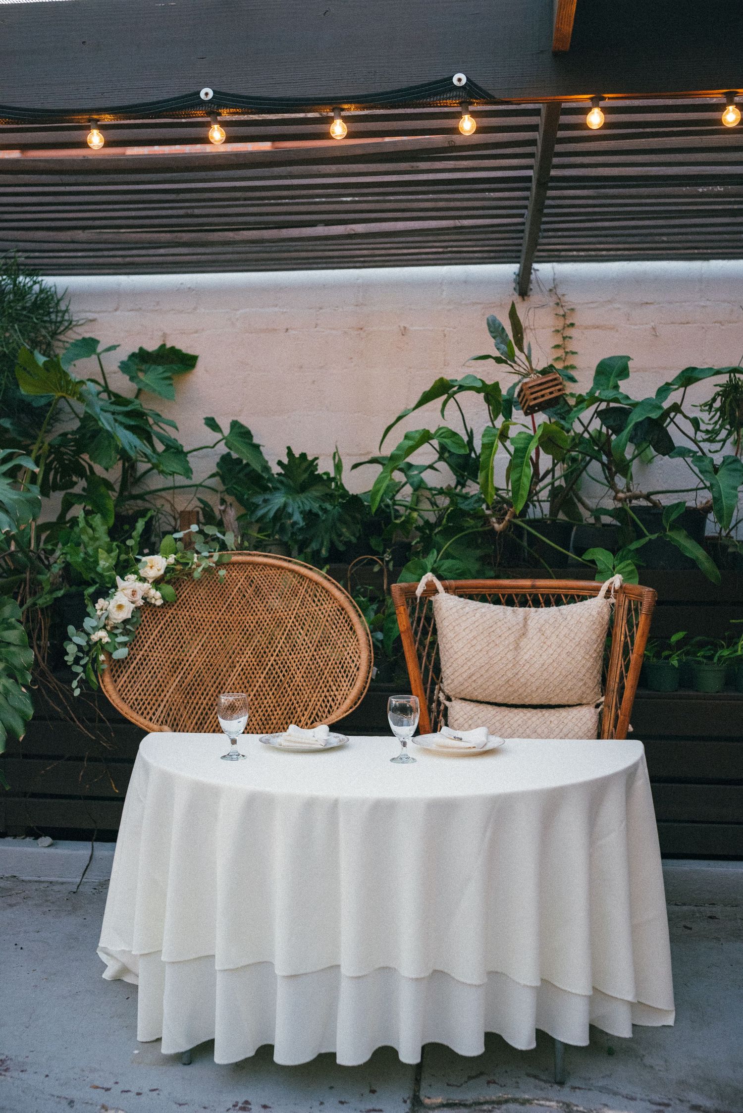 Intimate sweetheart table with wicker chairs and string lights for outdoor wedding reception.