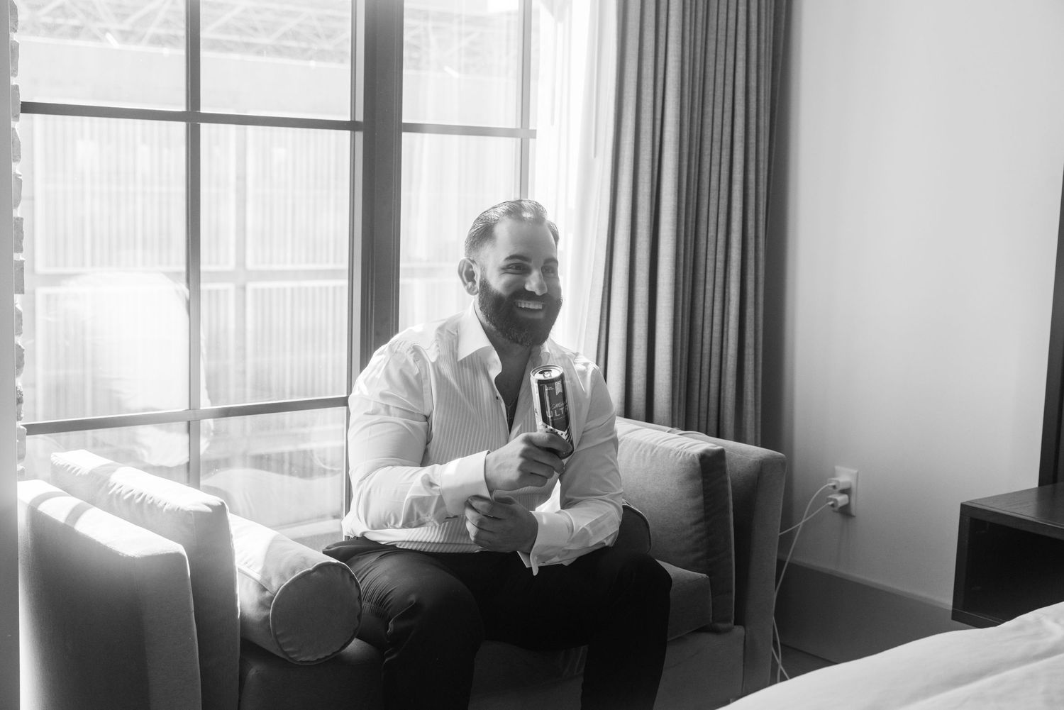 Man in white shirt smiling while sitting in armchair by window holding champagne glass in black and white photo.