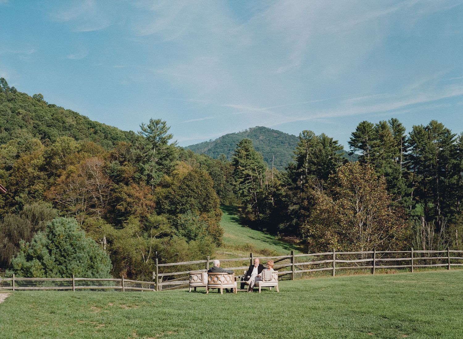Couple sits in Adirondack chairs on lush green lawn surrounded by mountains and trees at scenic outdoor venue.