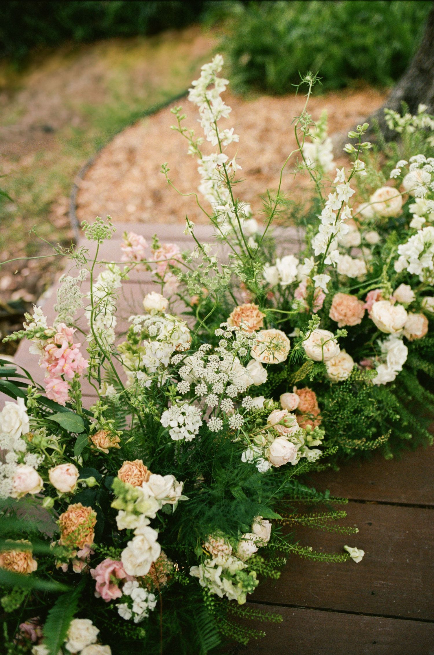 Delicate white and peach floral arrangements with greenery cascade along a rustic wooden surface in a garden setting.