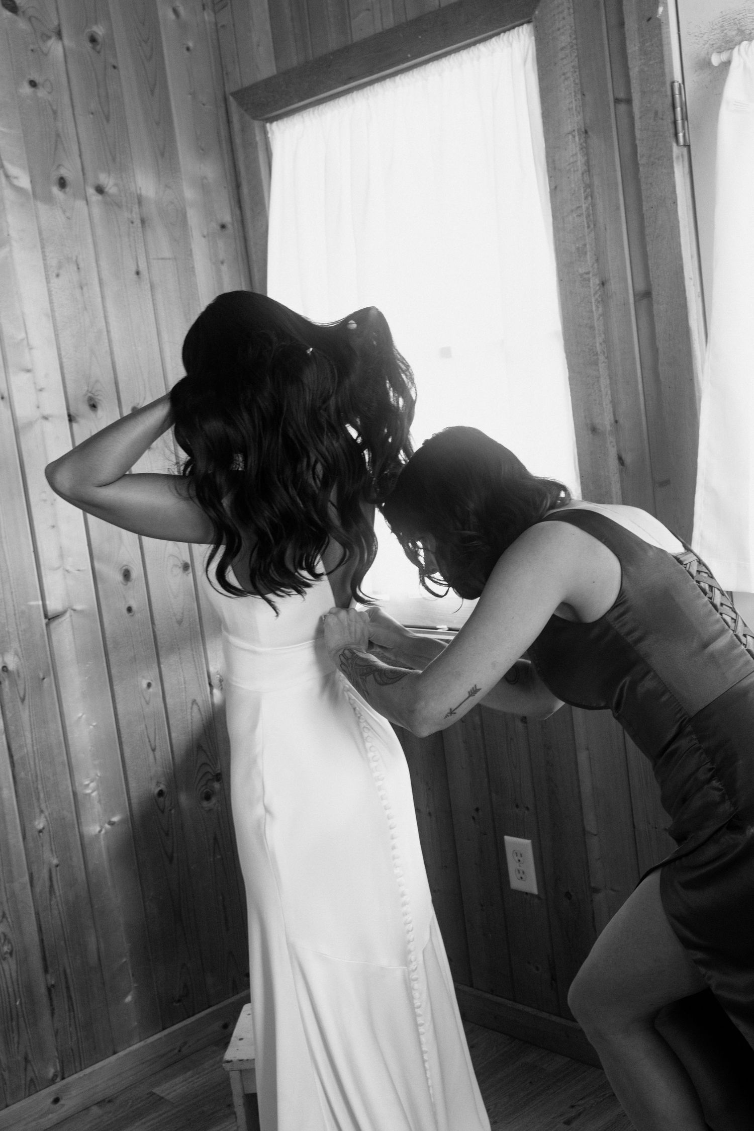 Black and white photo of bridesmaid buttoning the back of a brides elegant white wedding gown.