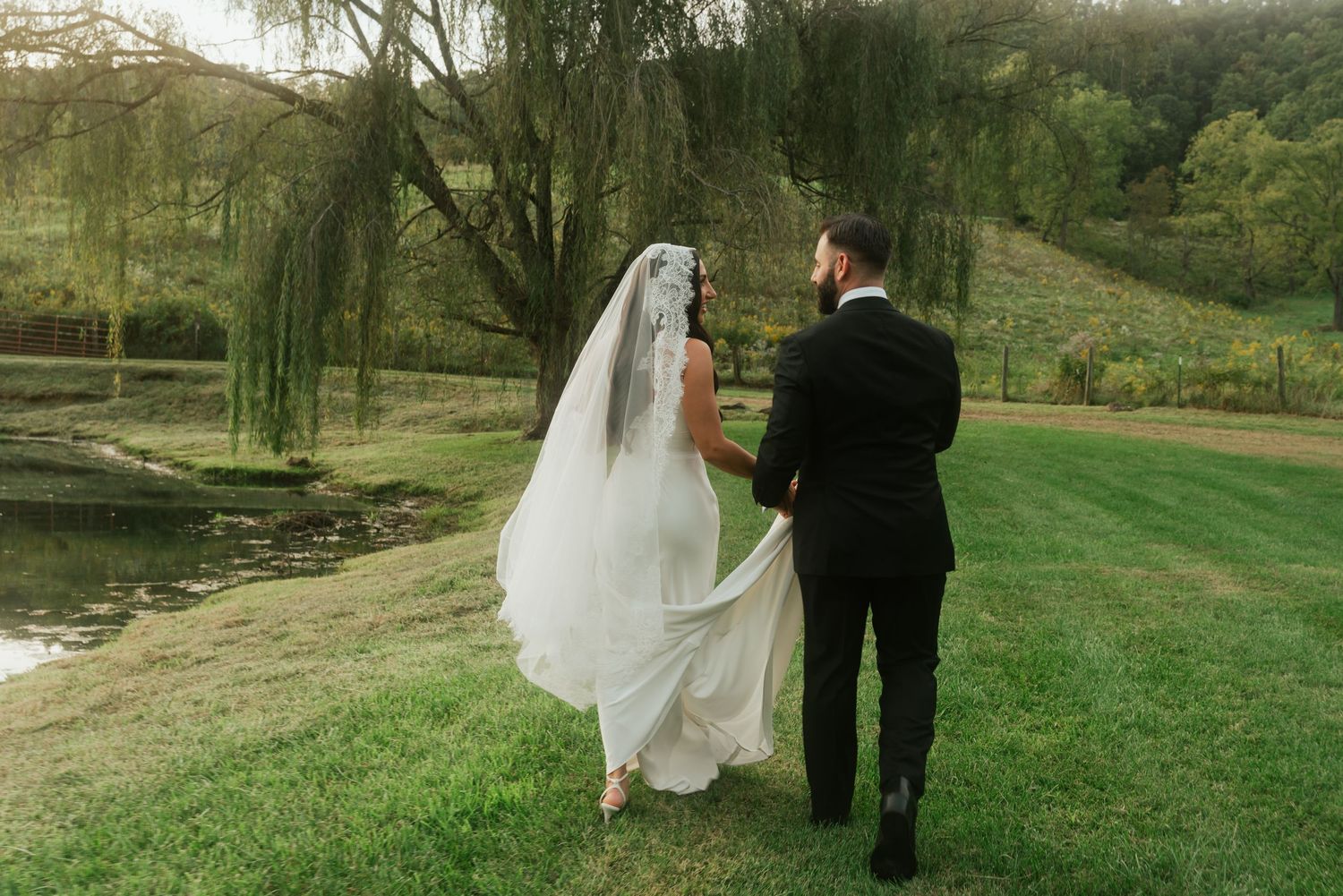 Bride and groom walking hand in hand on lush green grass near a pond, with a flowing veil and willow tree backdrop.
