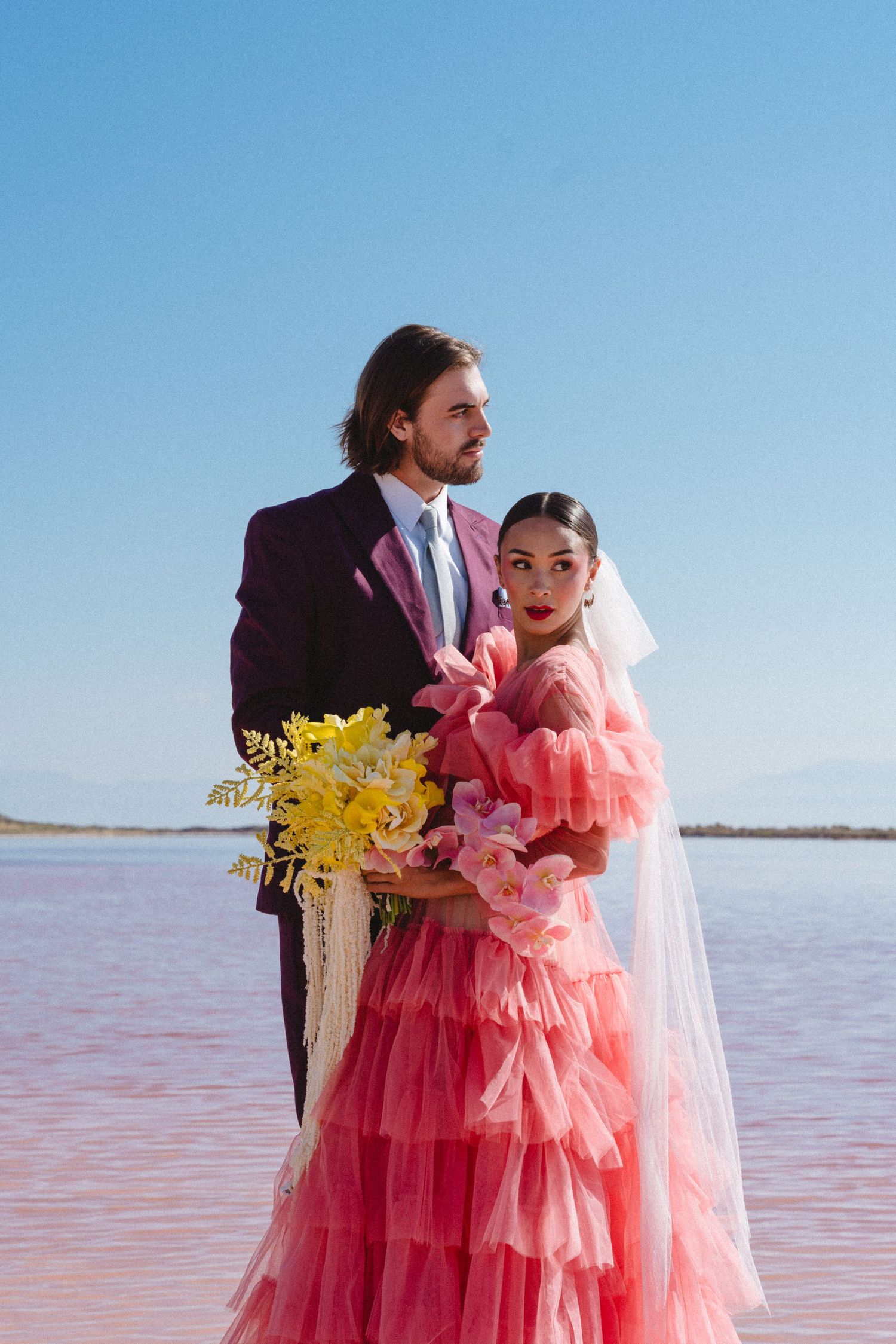 Wedding couple in pink dress and dark suit posing against pink salt lake backdrop.