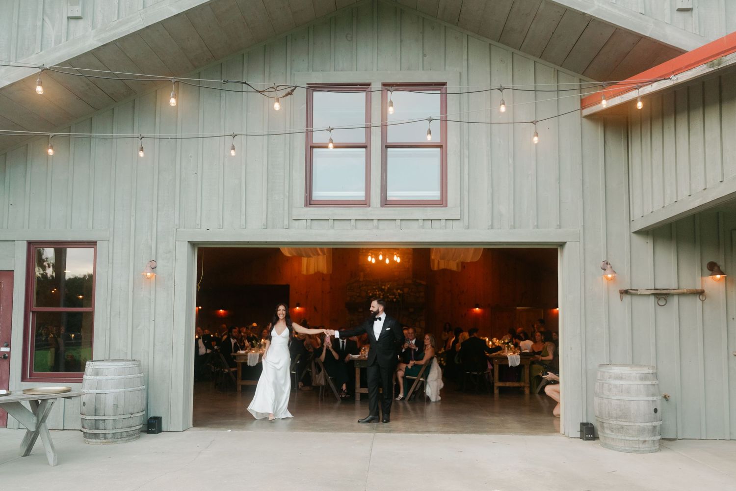Couple shares first dance outside rustic barn wedding venue with string lights and wine barrels.