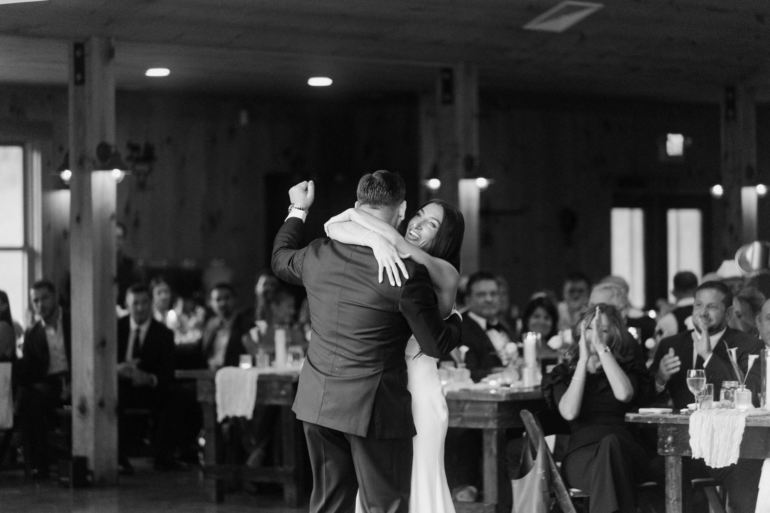 Bride and groom share their first dance in a rustic wedding venue while guests applaud in the background.