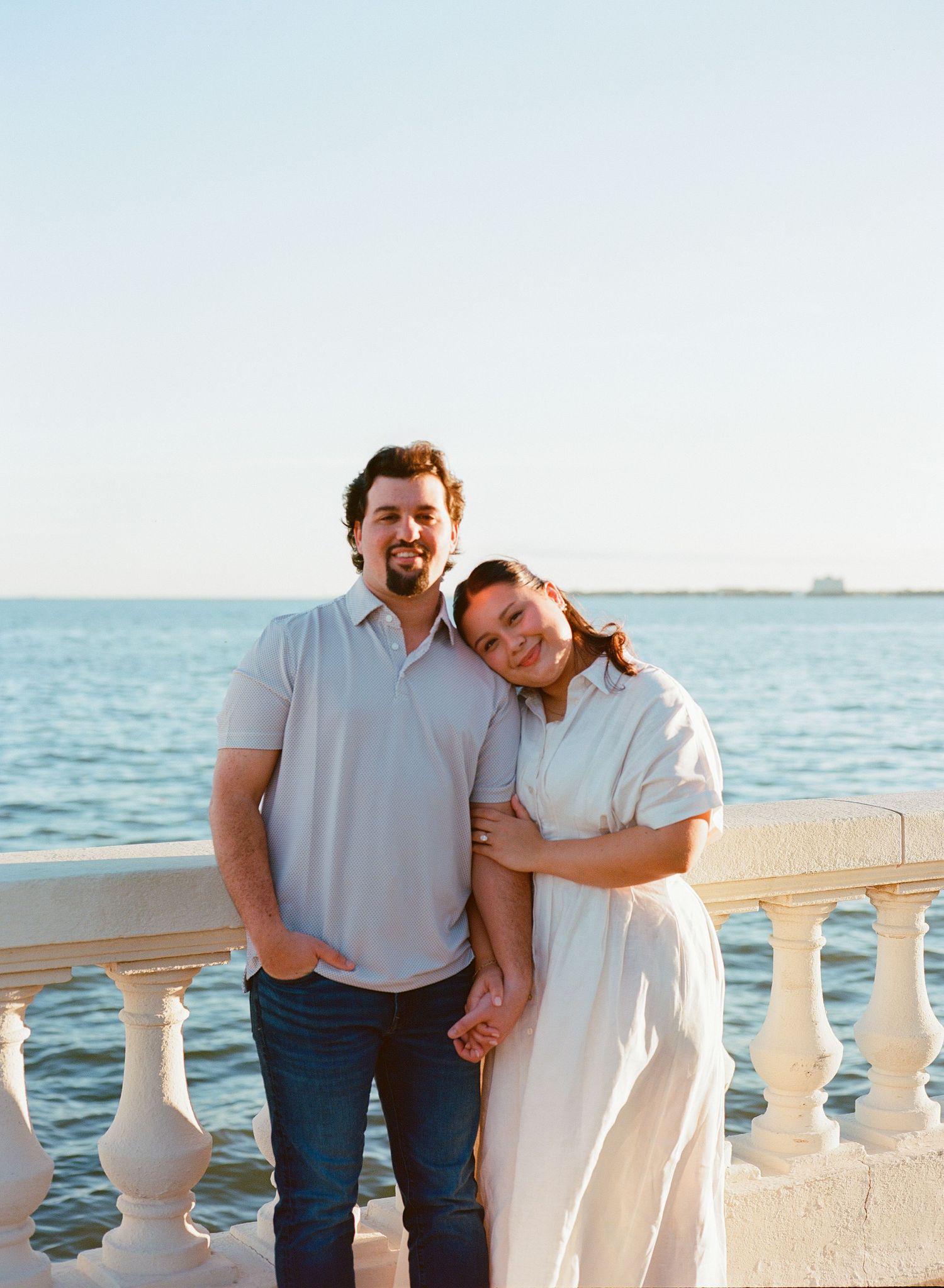 Couple smiling together on a waterfront terrace with white balustrade, woman leaning on man's shoulder at golden hour.