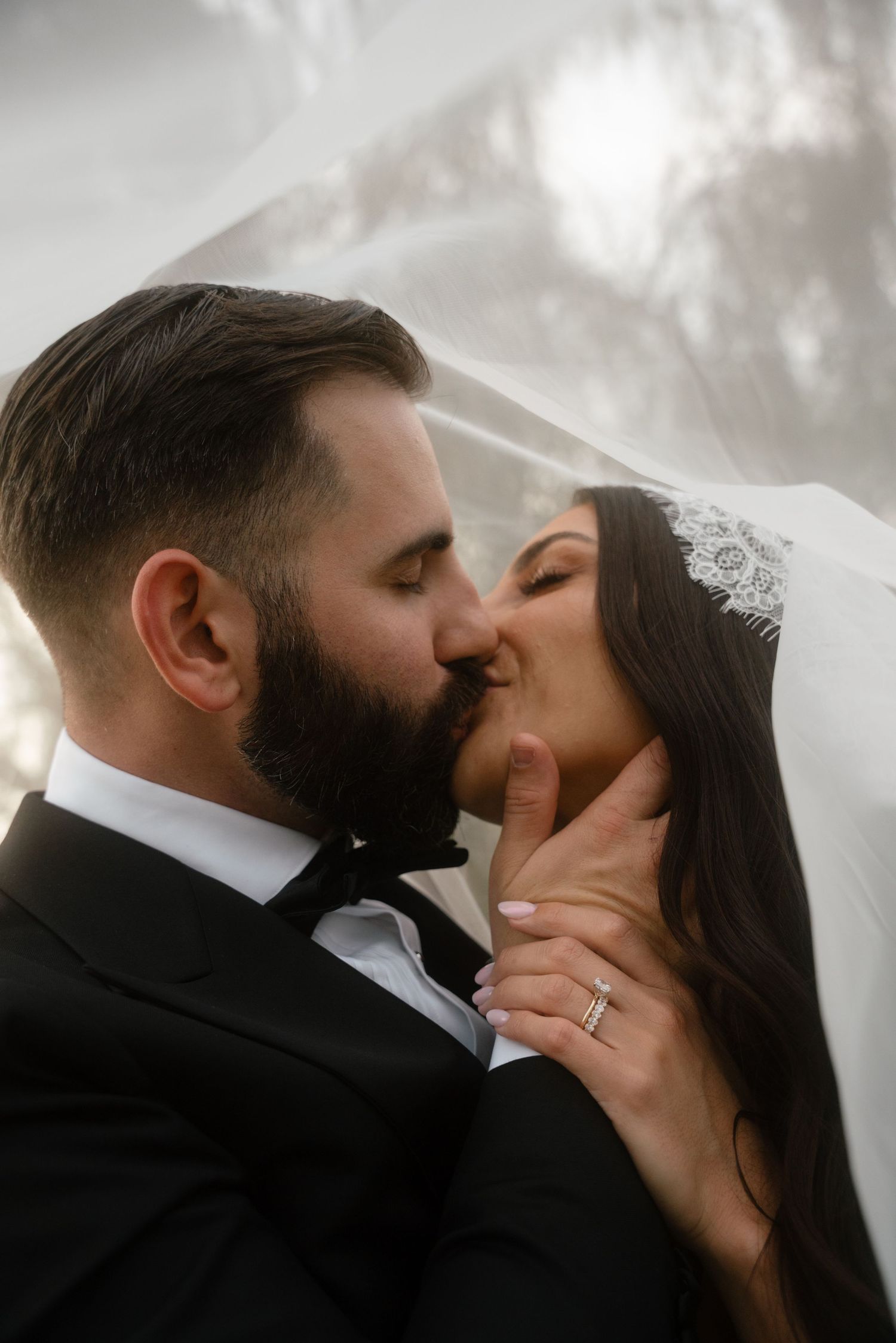 Bride and groom sharing a romantic kiss under a flowing veil on their wedding day.
