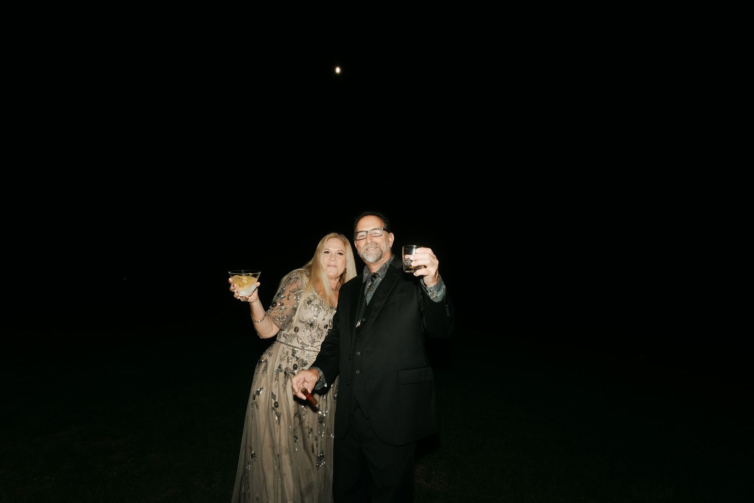 Couple smiling and taking a selfie outdoors at night, holding drinks under a moonlit sky.