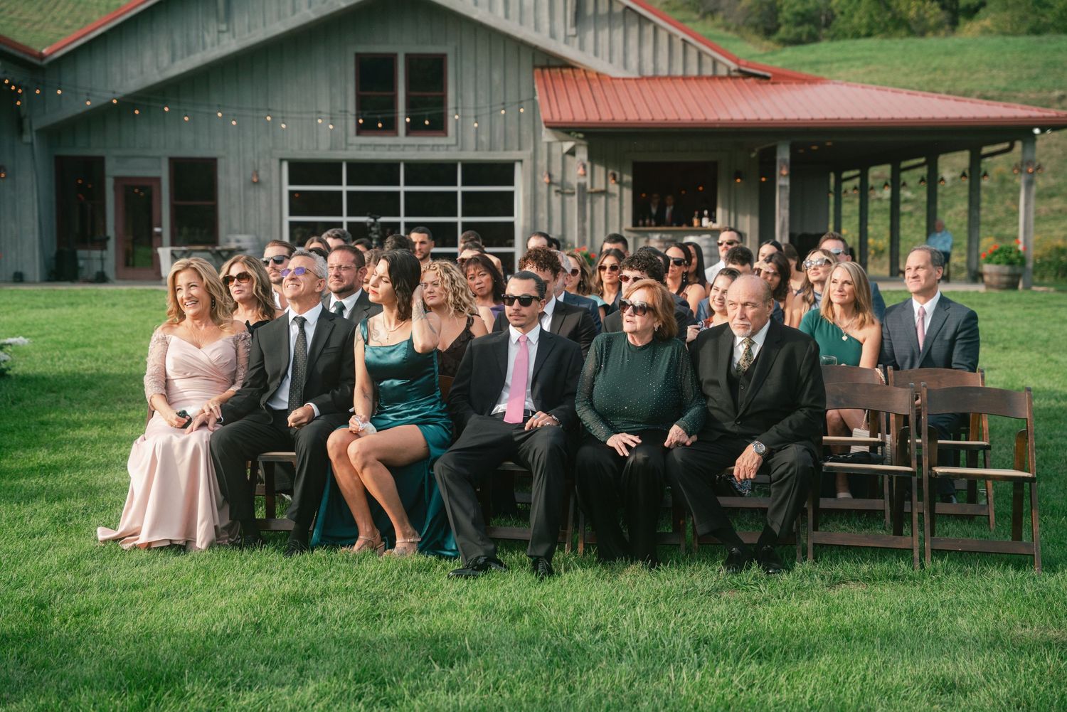 Wedding guests seated outdoors on wooden chairs at a rustic barn venue, dressed in formal attire for an outdoor ceremony.