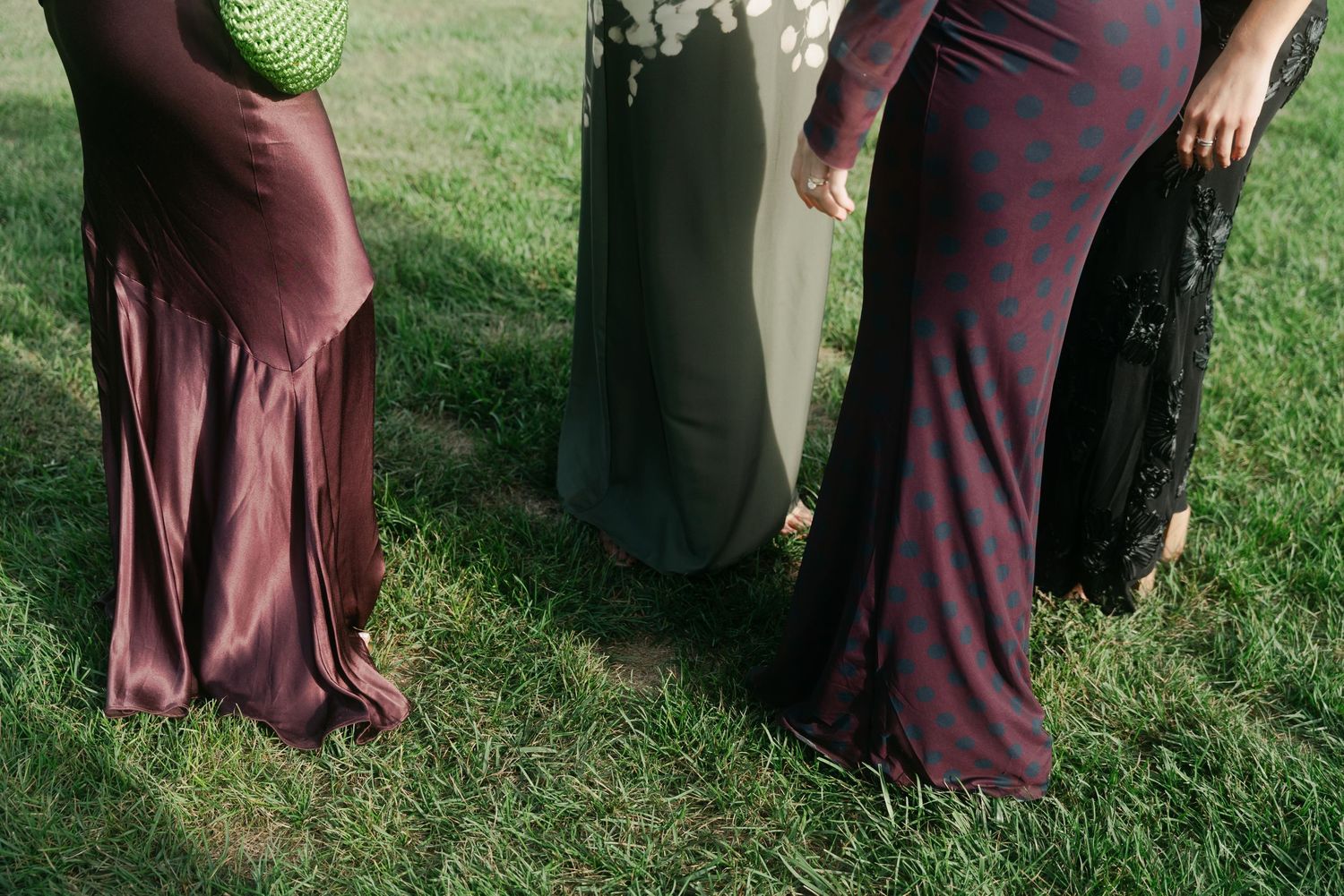Three women wearing long maxi dresses standing on green grass, showing elegant floor-length gowns in mauve, gray, and burgundy.