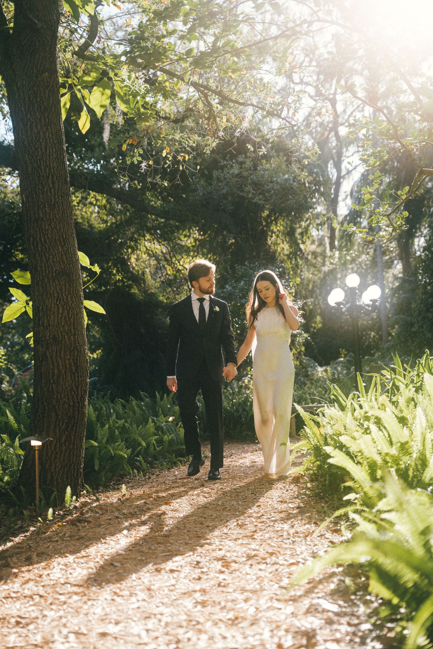 Sun-drenched wedding photos of couple walking through a tree-lined garden path.