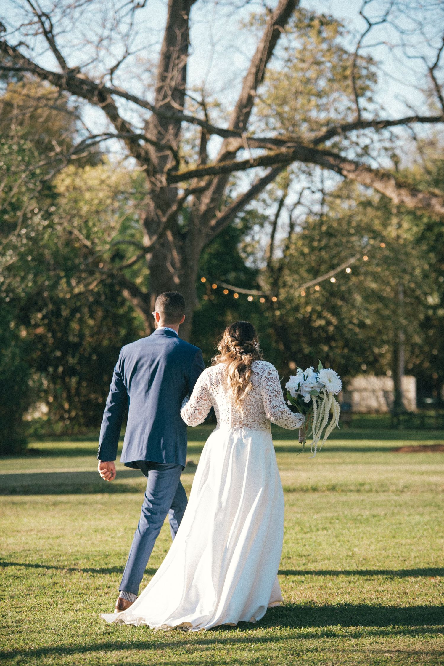 A couple walks hand in hand across a sunlit lawn with string lights hanging in the trees above.