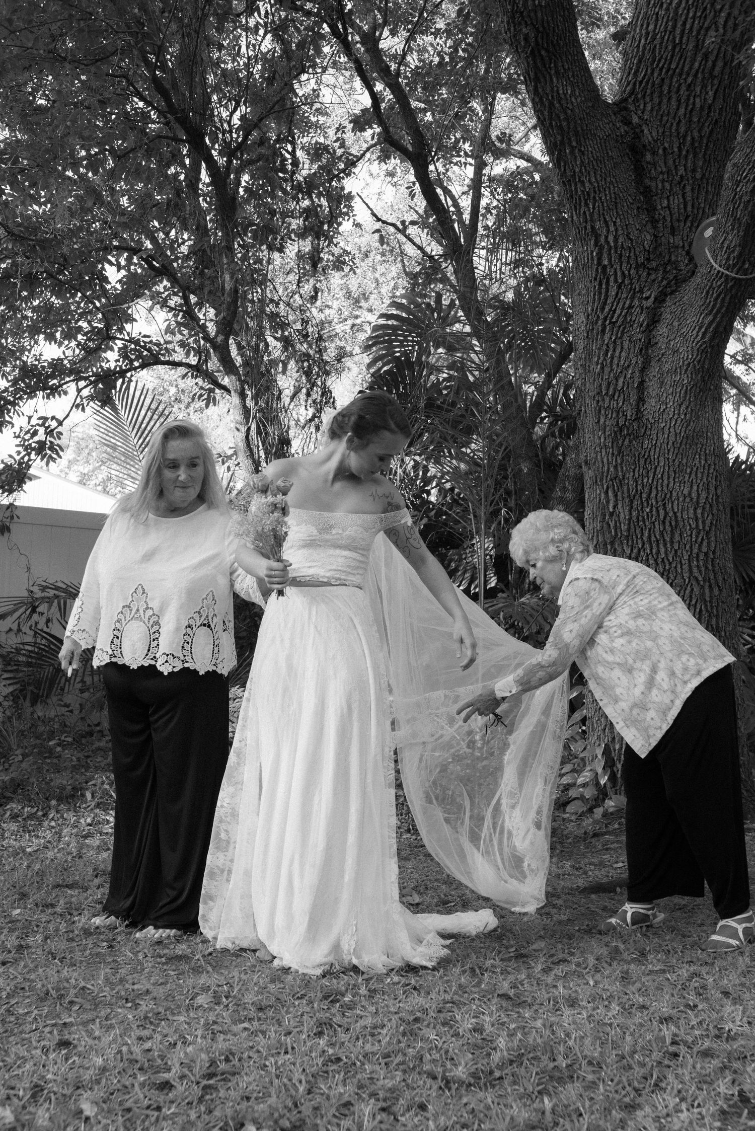 Wedding party helping with bridal preparations outdoors under large trees.