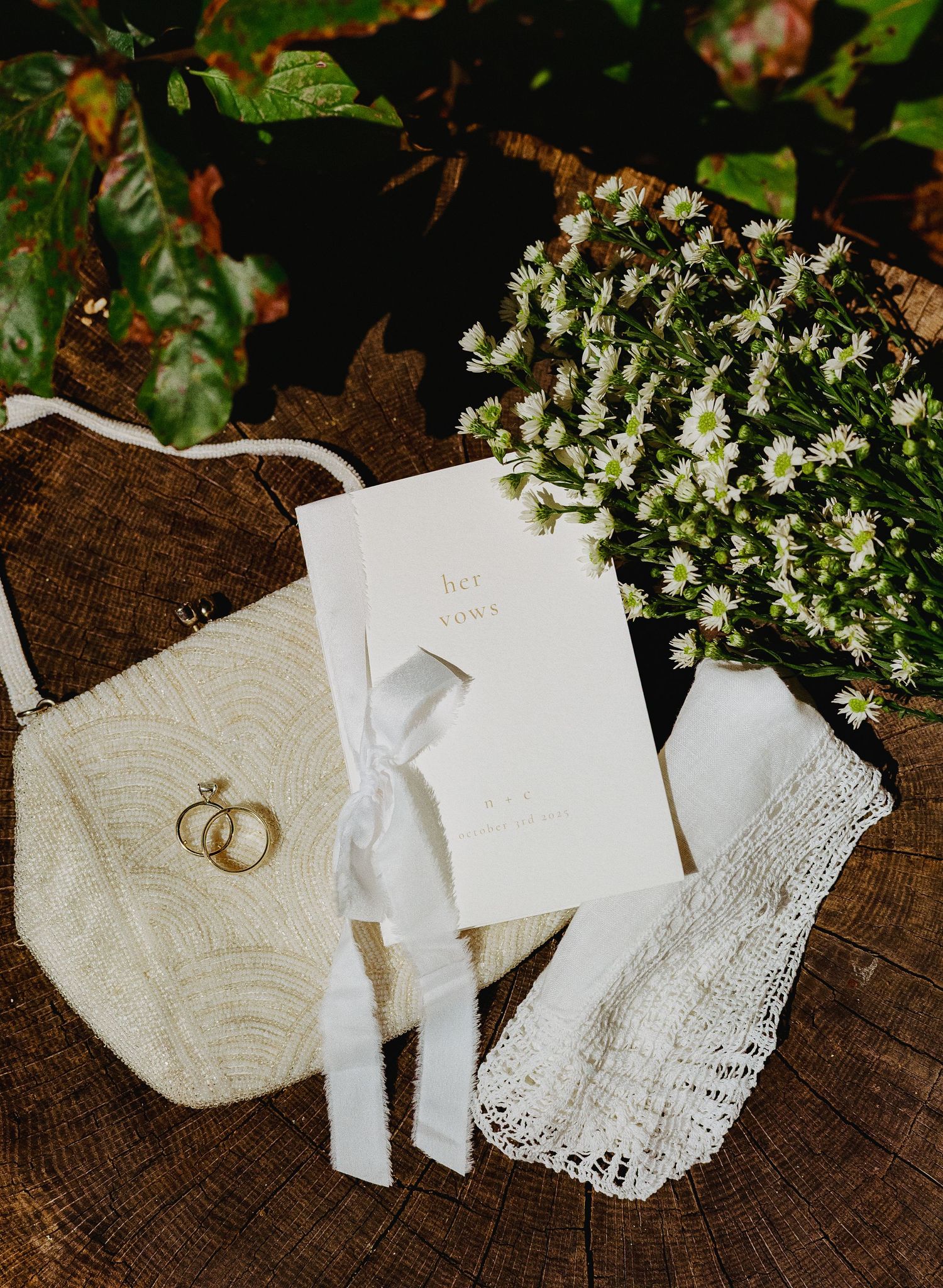 Wedding flat lay with gold rings, white floral bouquet, bridal purse, vows booklet, and lace handkerchief on rustic wood.