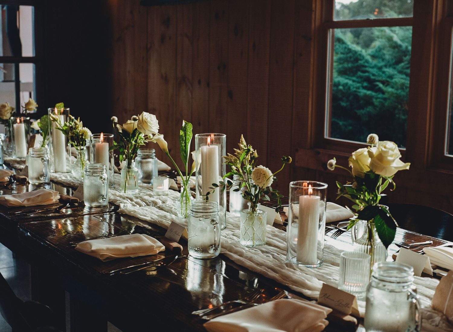 Elegant wedding reception table with white candles, roses, and greenery centerpieces on a dark wood table.