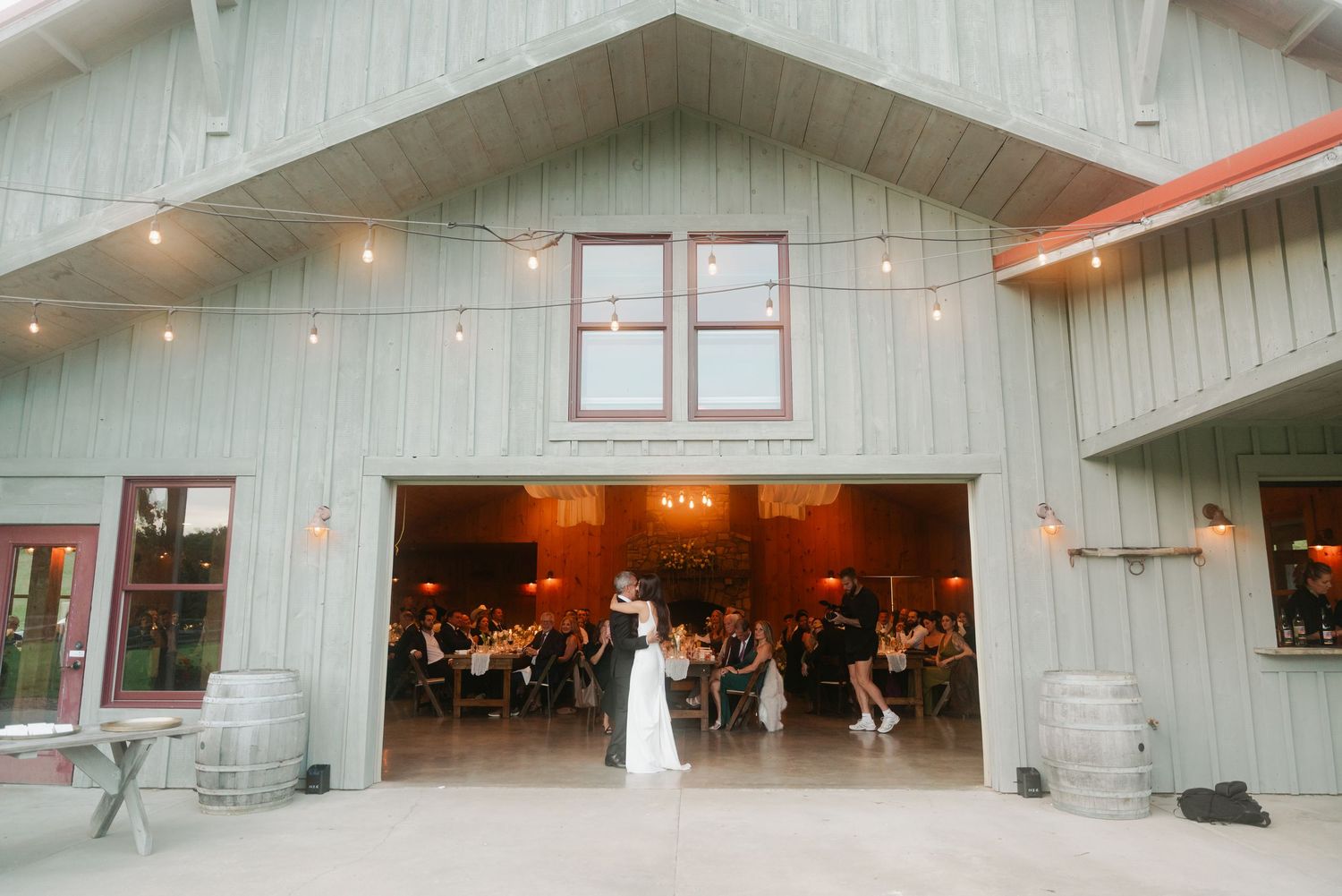Couple shares first dance outside rustic barn wedding venue with string lights and guests dining inside.