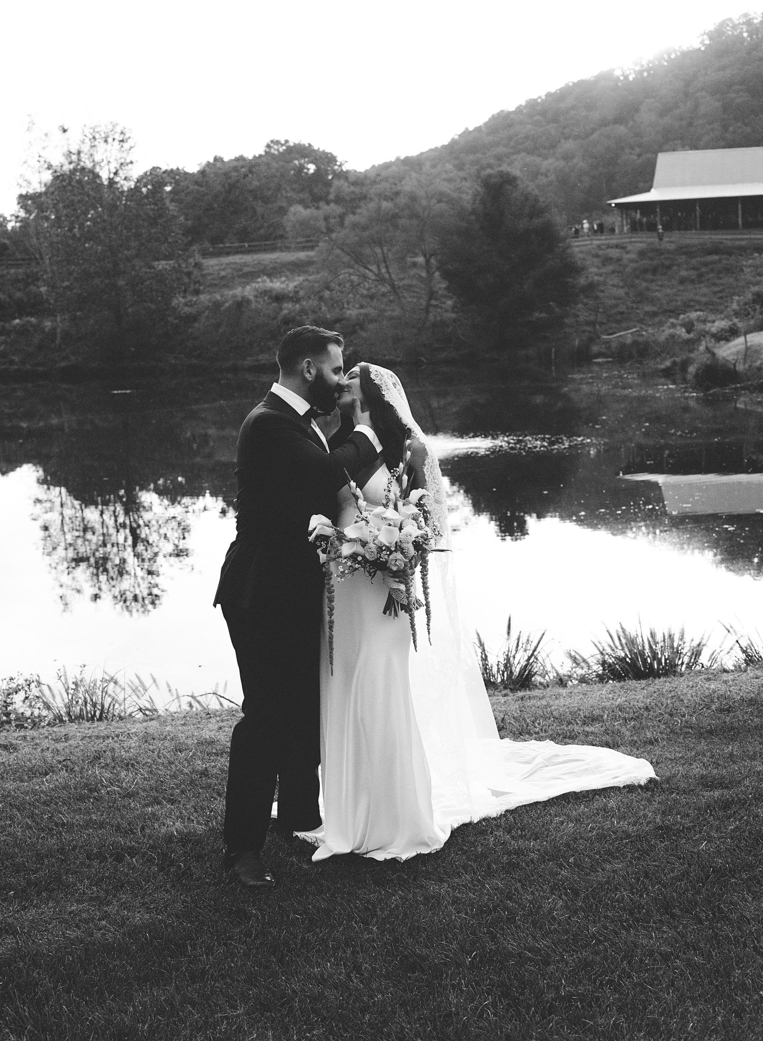 Black and white photo of bride and groom kissing by a pond with rolling hills and a barn in the background.