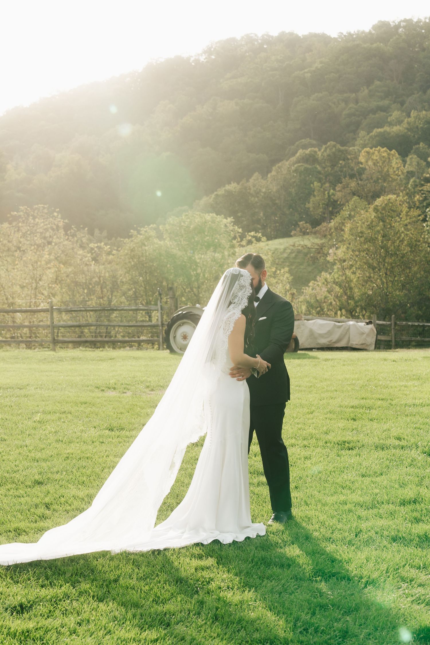 Bride and groom sharing a romantic moment on a sunlit green lawn with a scenic mountain backdrop on their wedding day.