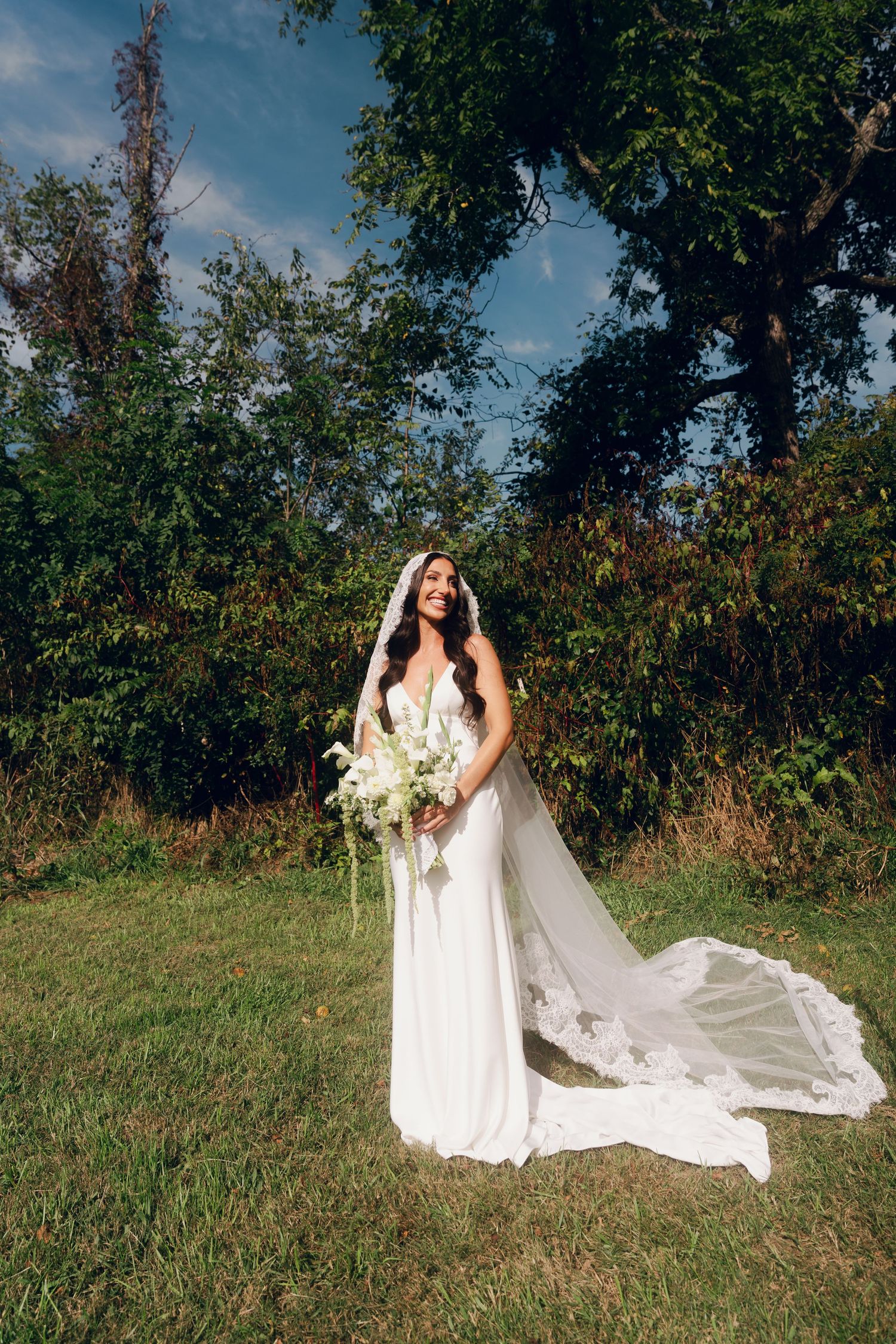 Bride in white gown and veil holds white bouquet outdoors on green grass with trees and blue sky.
