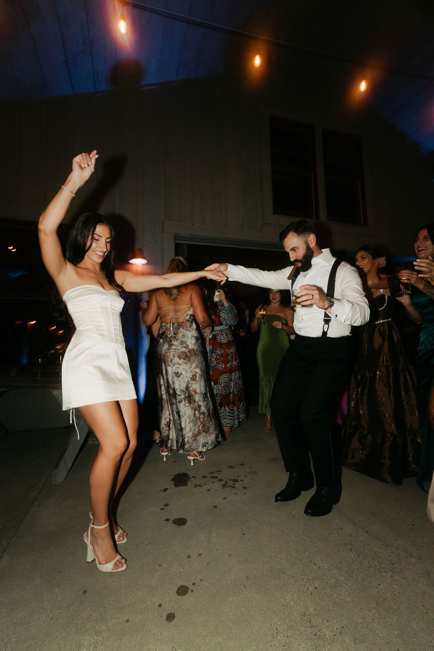 Bride in white mini dress dancing with groom in suspenders at outdoor wedding reception at night.
