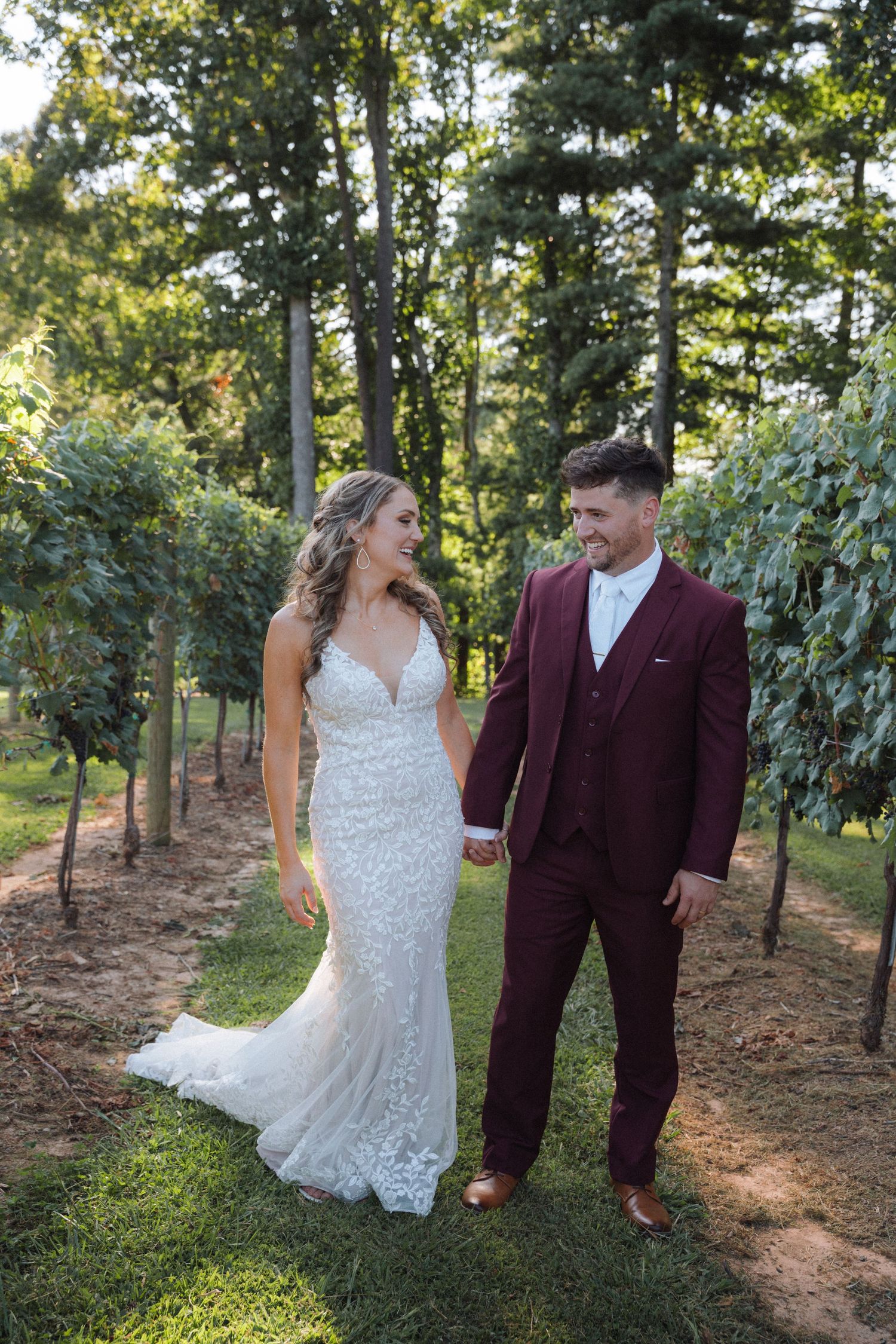 A couple shares joyful moments together in a vineyard setting wearing burgundy and white wedding attire.