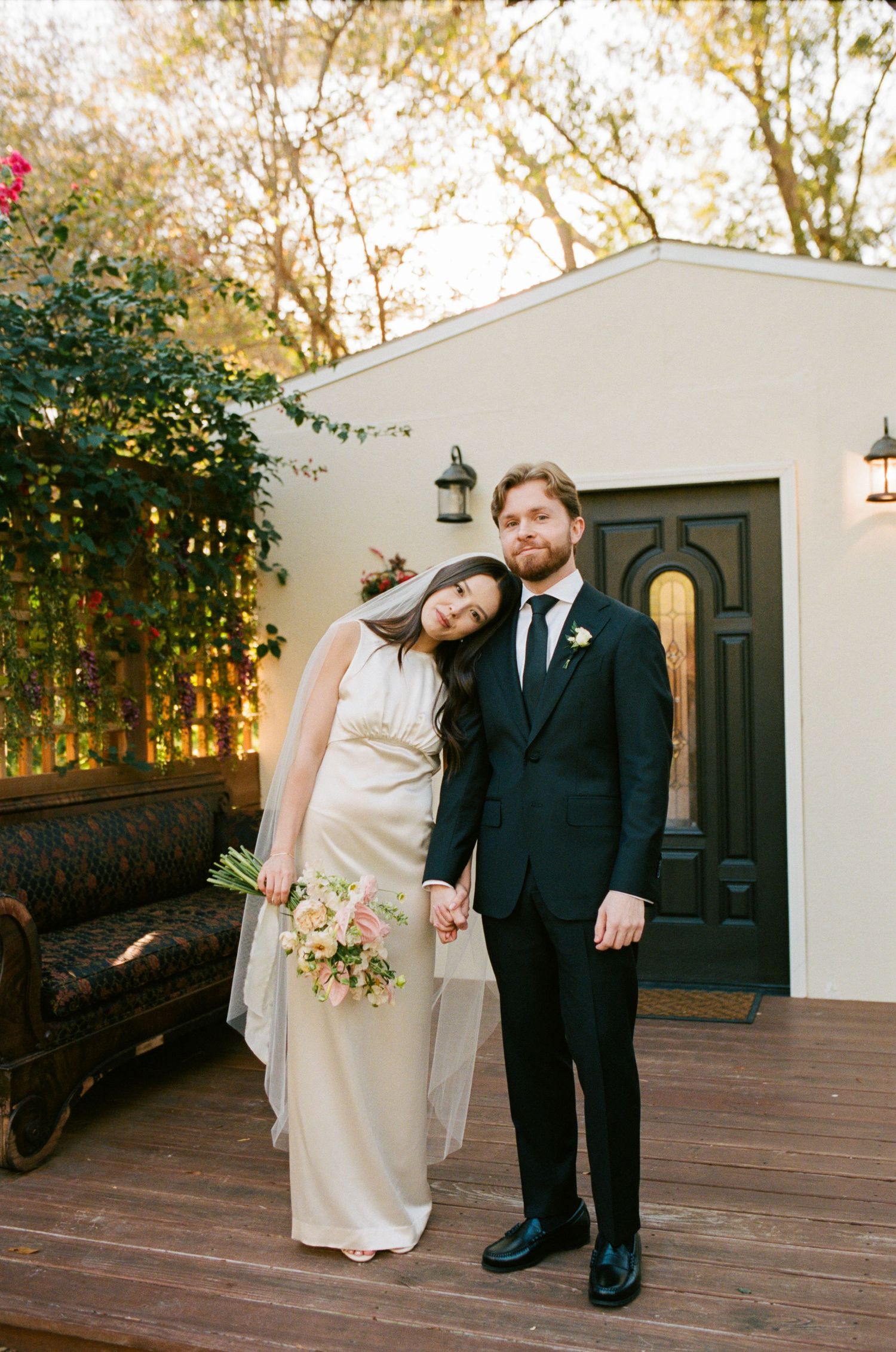 A couple poses in front of a white building with a dark wooden door at sunset.