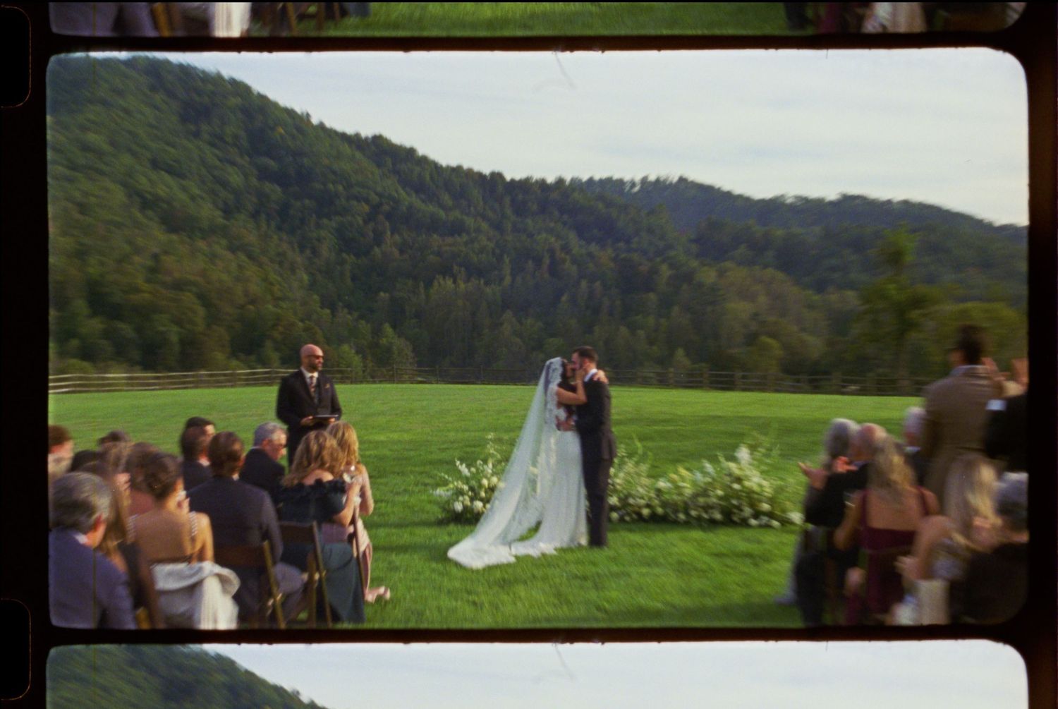 Wedding guests gather on a grassy hillside with mountain vistas in the background.