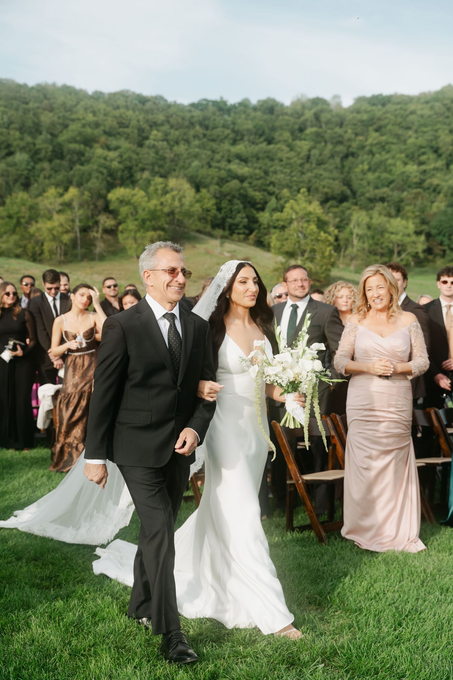 Bride in white gown walks down outdoor aisle with father, guests seated on green hillside backdrop.