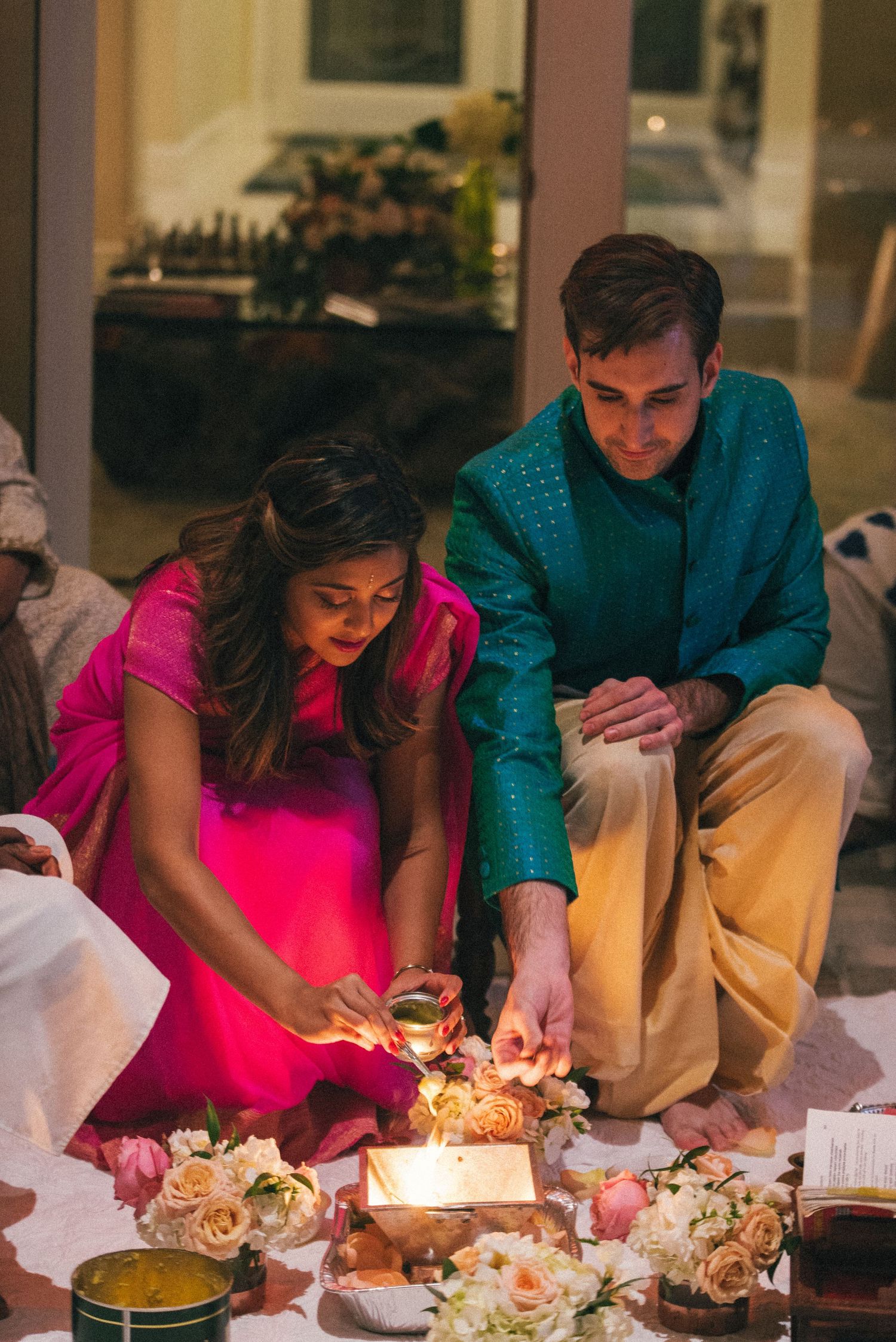 A couple in vibrant pink and teal outfits light candles at an elegantly decorated table with flowers.