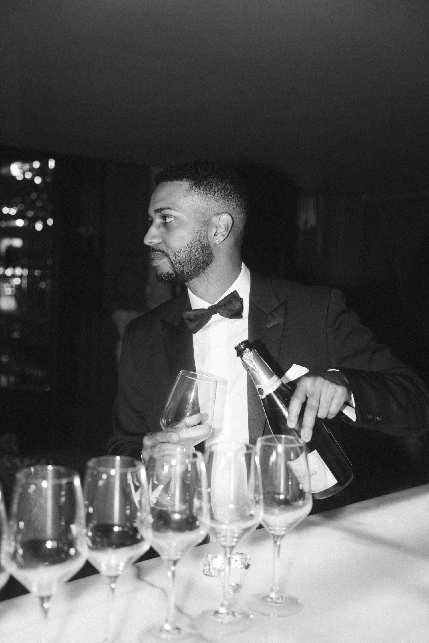 Elegant black and white photo of a groom in a tuxedo and bowtie seated at a formal dinner table with wine glasses.