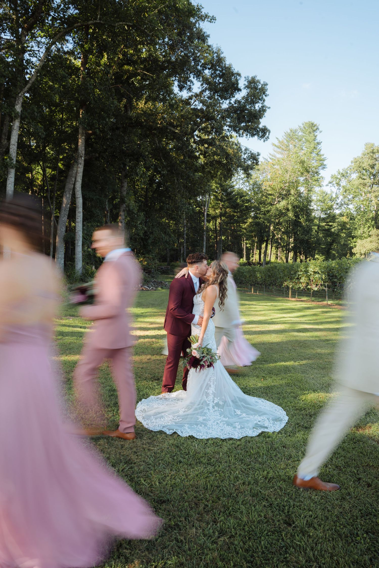 Wedding party in pink dresses and suits moving through a sunlit garden setting.