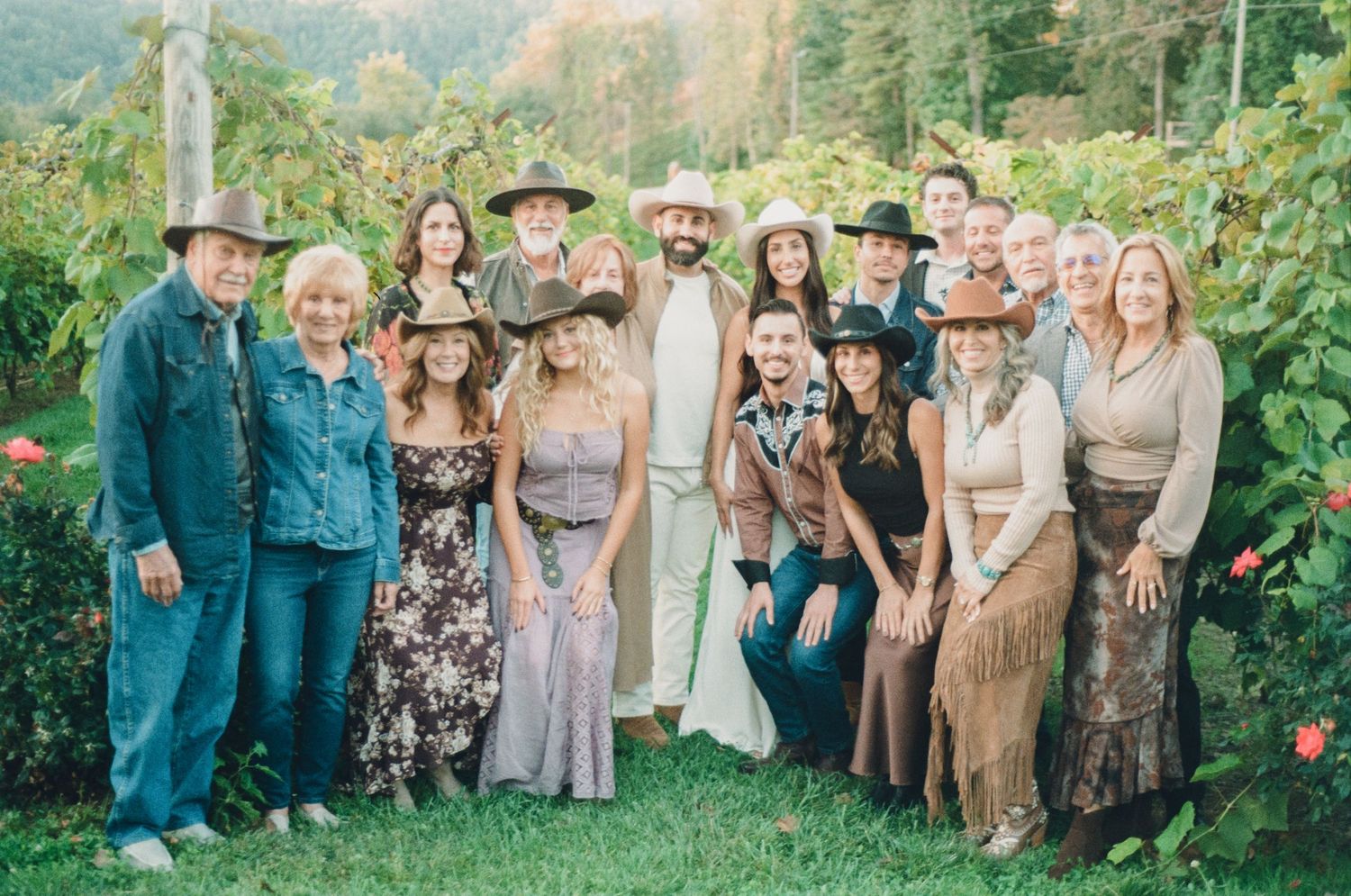 Large group of people posing together in a vineyard, wearing western-style outfits including cowboy hats and fringe.