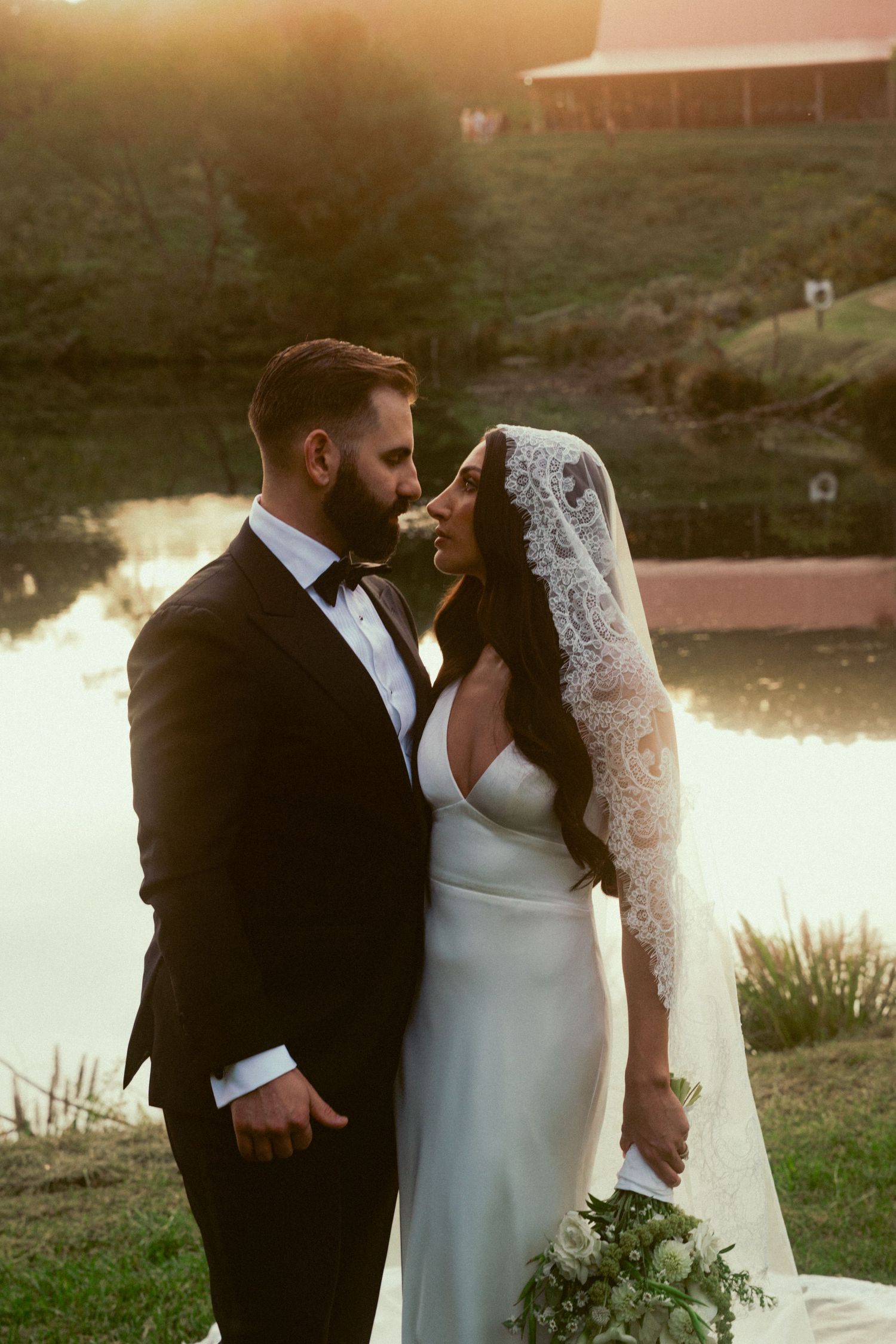 Bride and groom share a romantic moment at golden hour beside a pond, she wearing a lace veil and white gown.