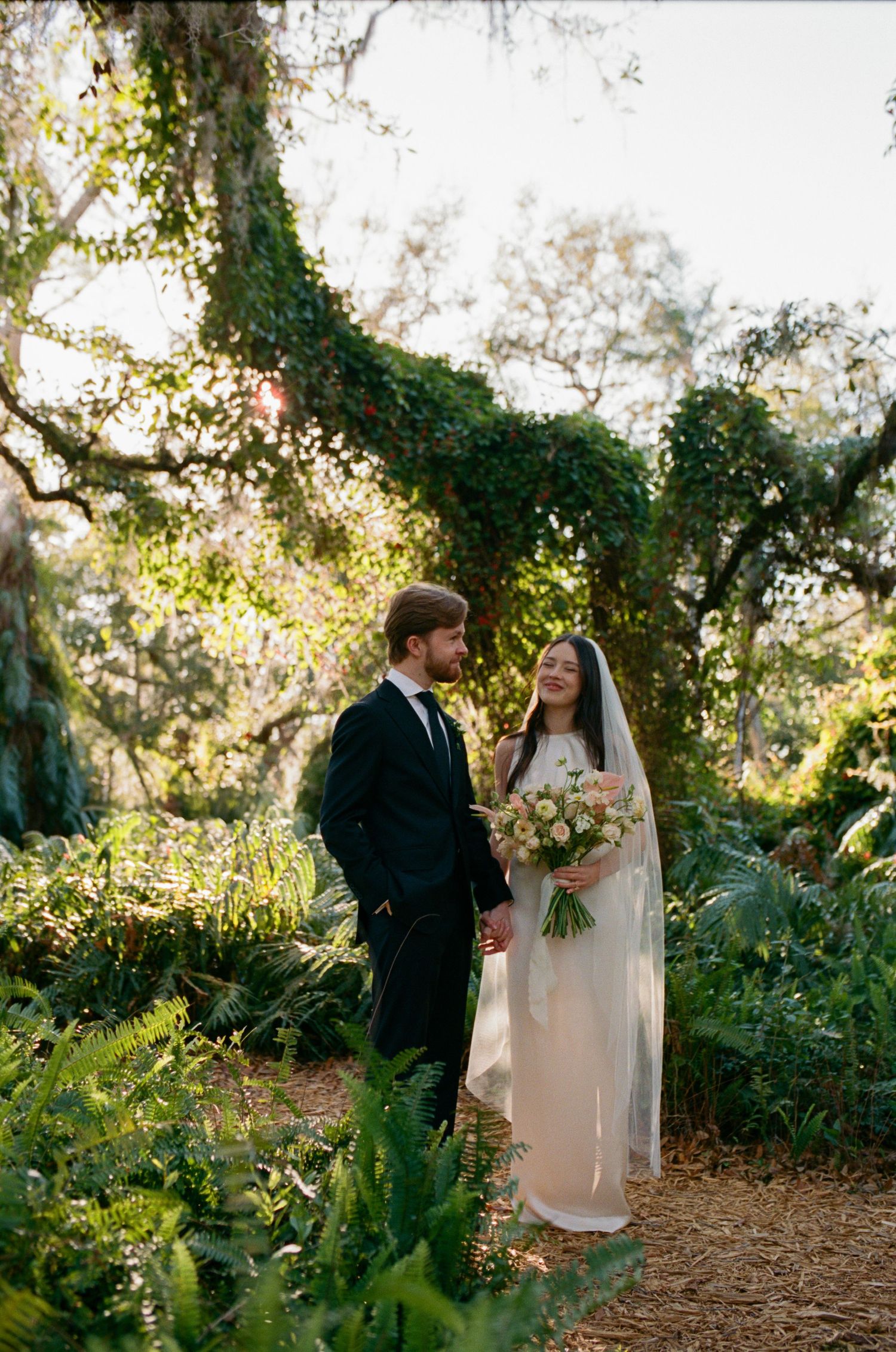 A wedding couple stands in a lush garden setting with ivy-covered trees.
