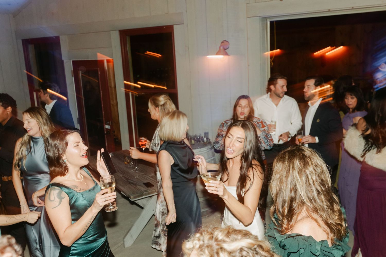 Wedding guests socializing and dancing with drinks at lively indoor reception celebration.