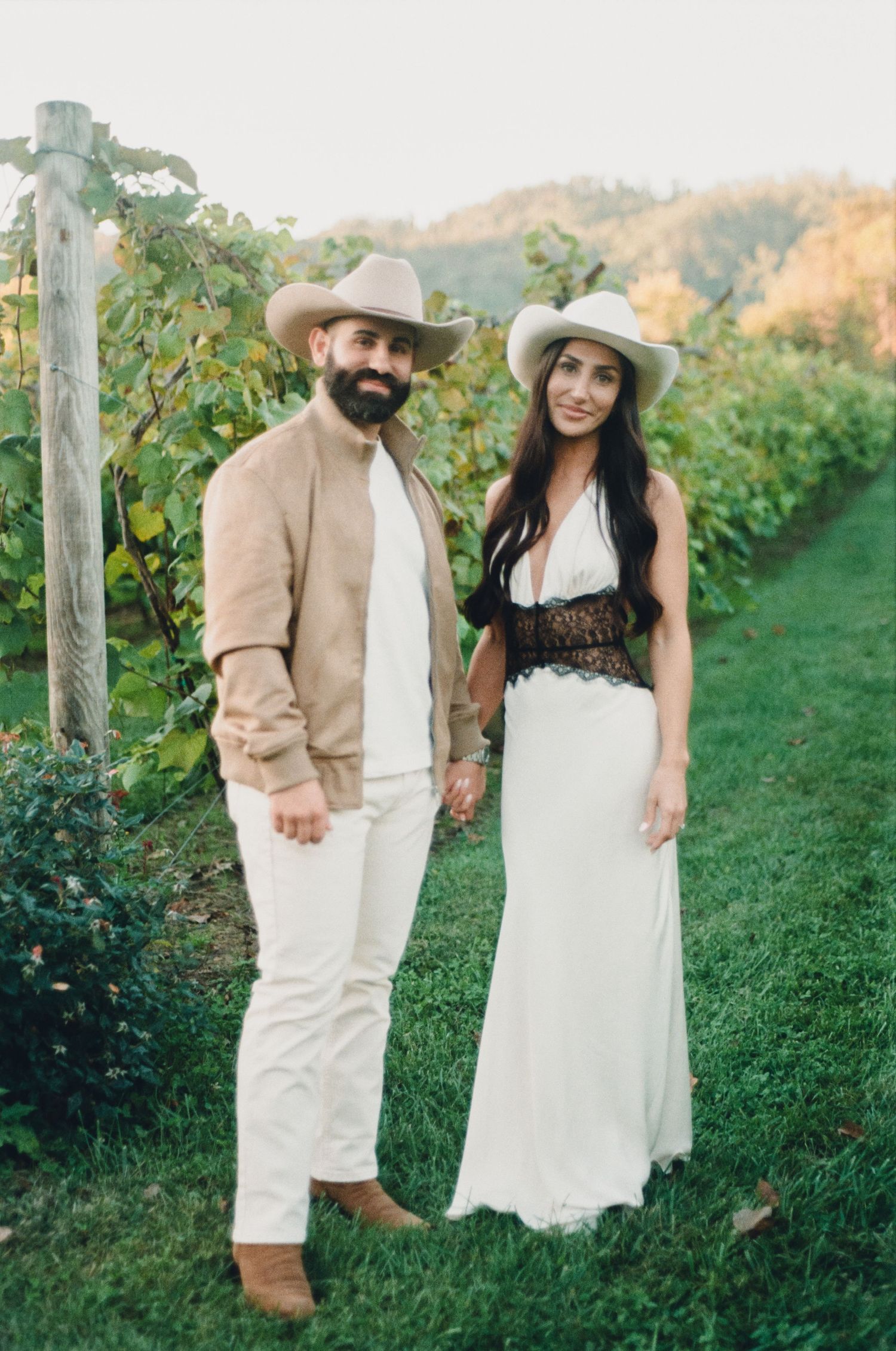 A bearded man and woman in cowboy hats and elegant attire pose together in a lush vineyard at golden hour.