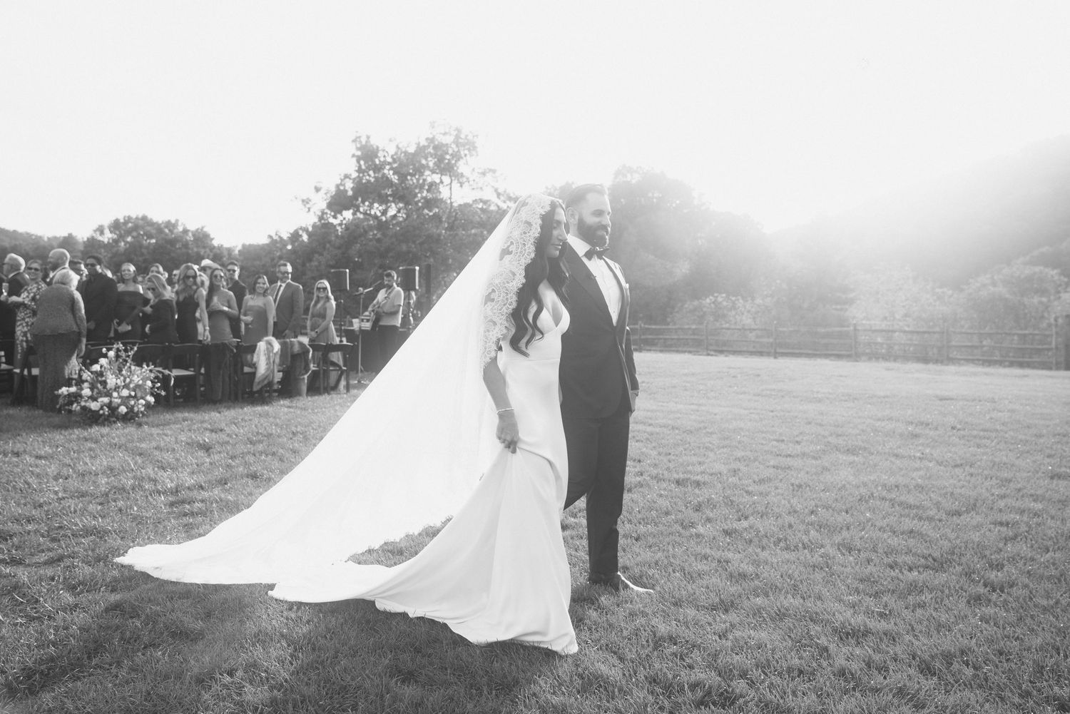 Bride and groom pose on green lawn after outdoor wedding ceremony, veil flowing in the breeze, black and white.