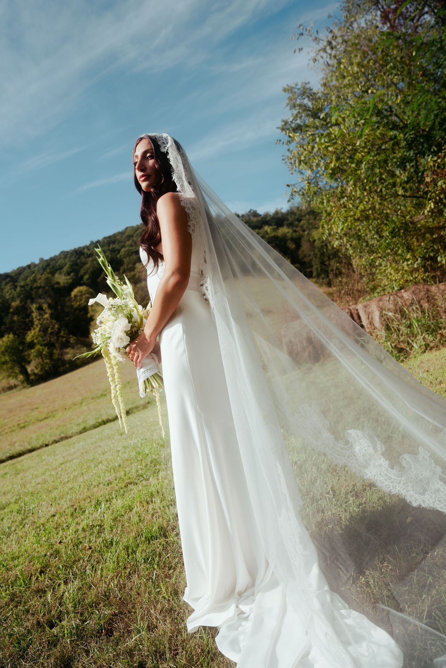 Bride in white gown and long veil holds bouquet outdoors in green field with blue sky and trees.