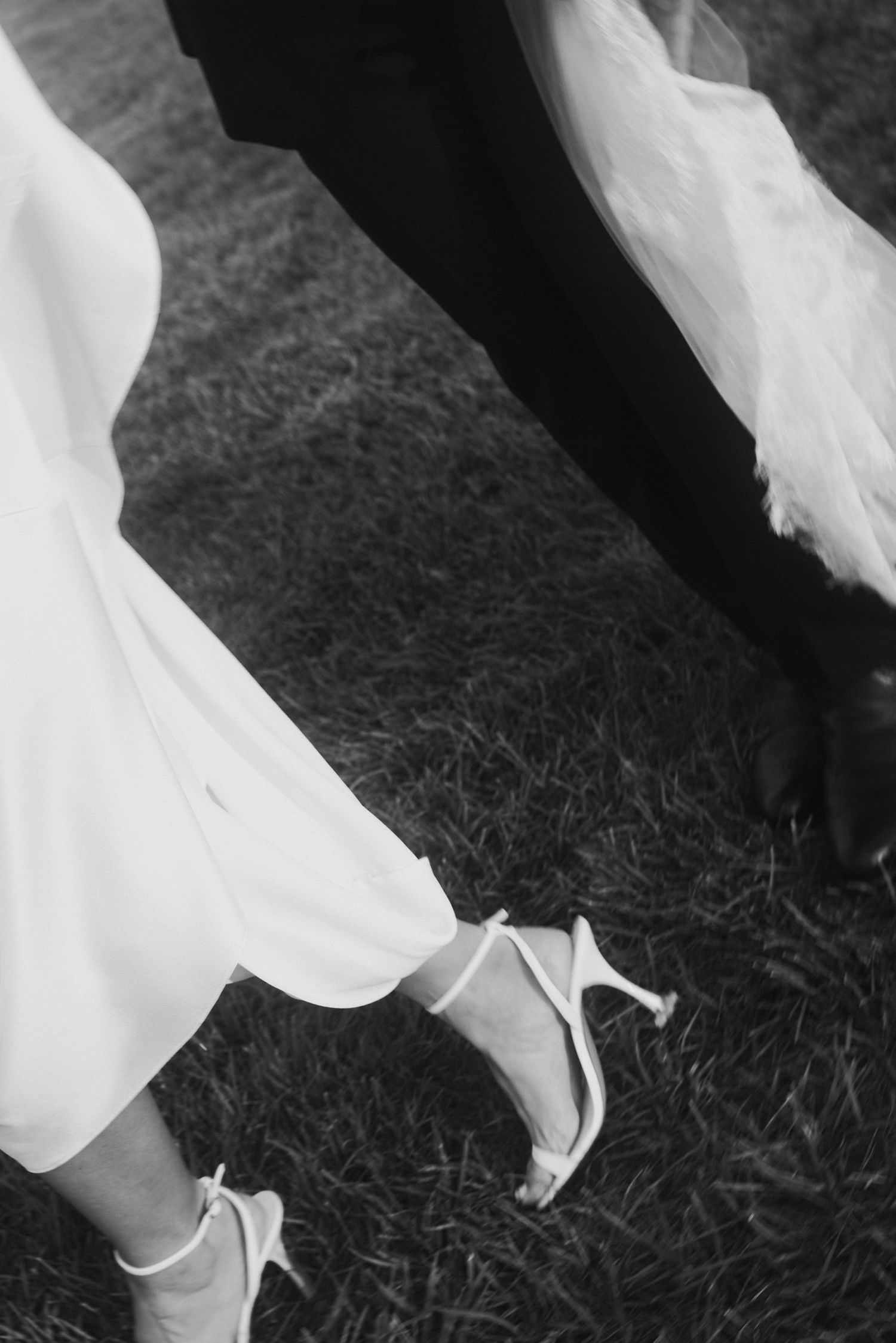 Black and white detail shot of a bride's white heels and flowing dress on grass.