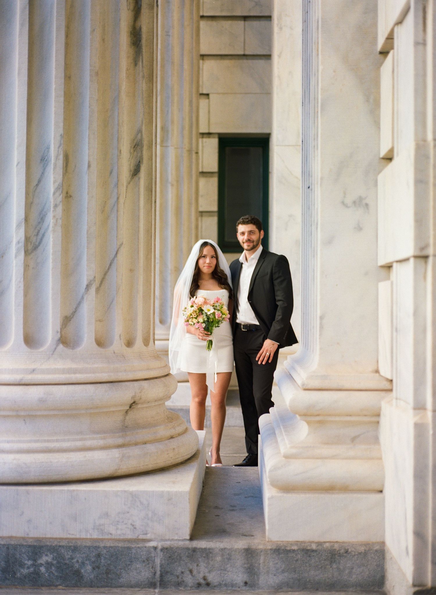 Bride in short white dress with veil and groom in black suit walk between marble columns holding bouquet.