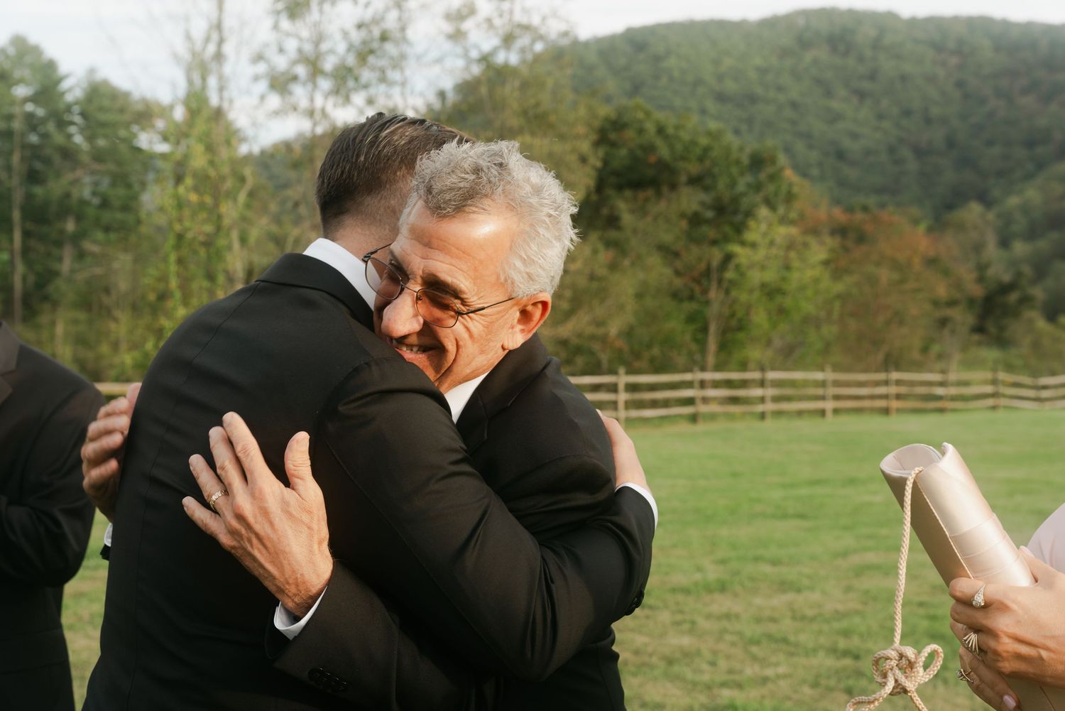 Two men in suits share a warm embrace outdoors at a wedding ceremony with green hills in the background.