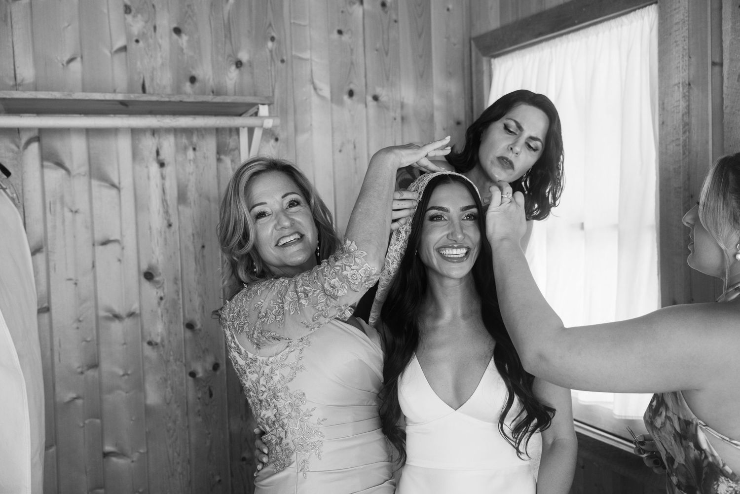 Bride smiling with bridesmaids helping style her hair in a rustic wooden room during wedding preparations.
