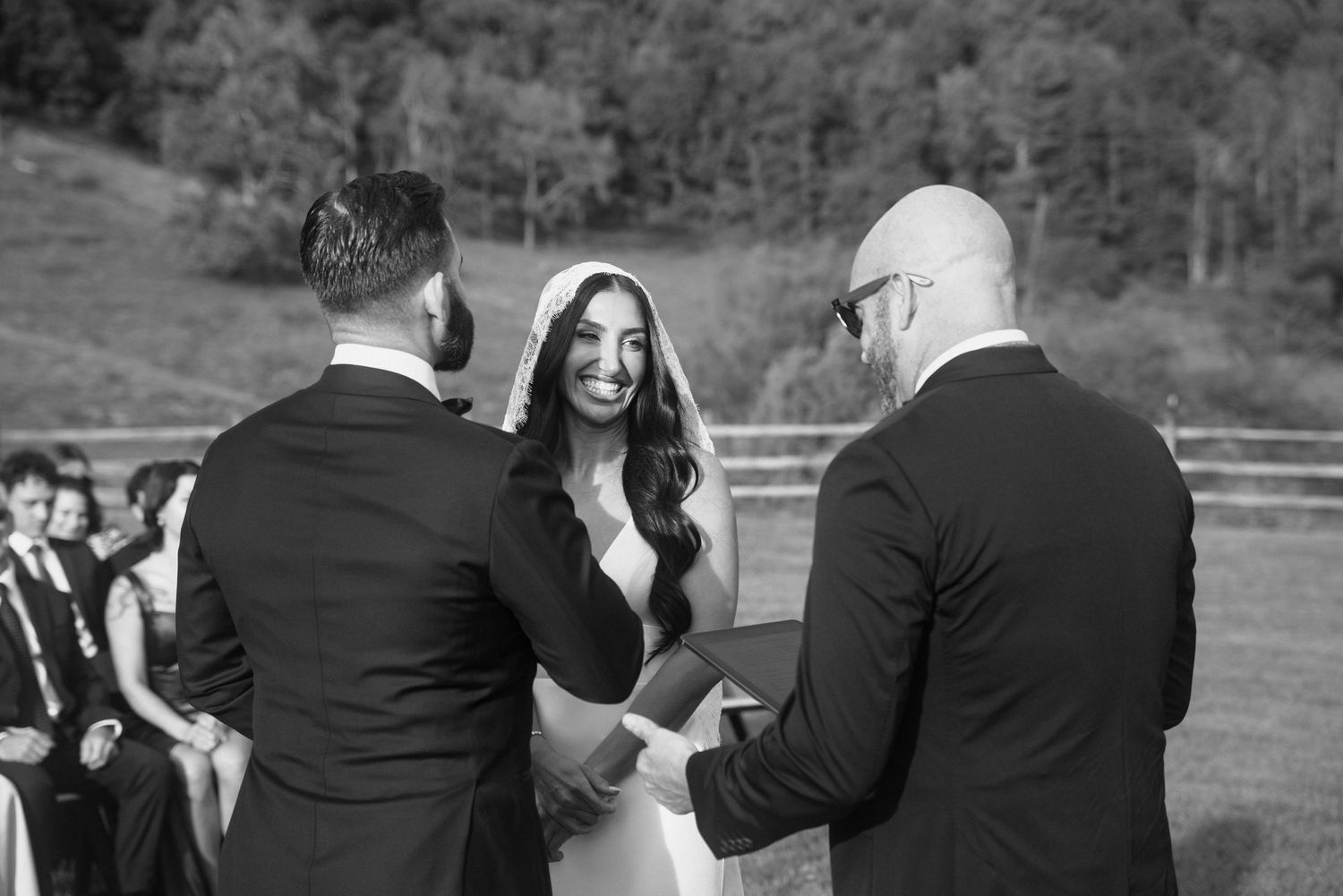 Bride smiling joyfully during outdoor wedding ceremony while exchanging vows with groom, black and white photo.