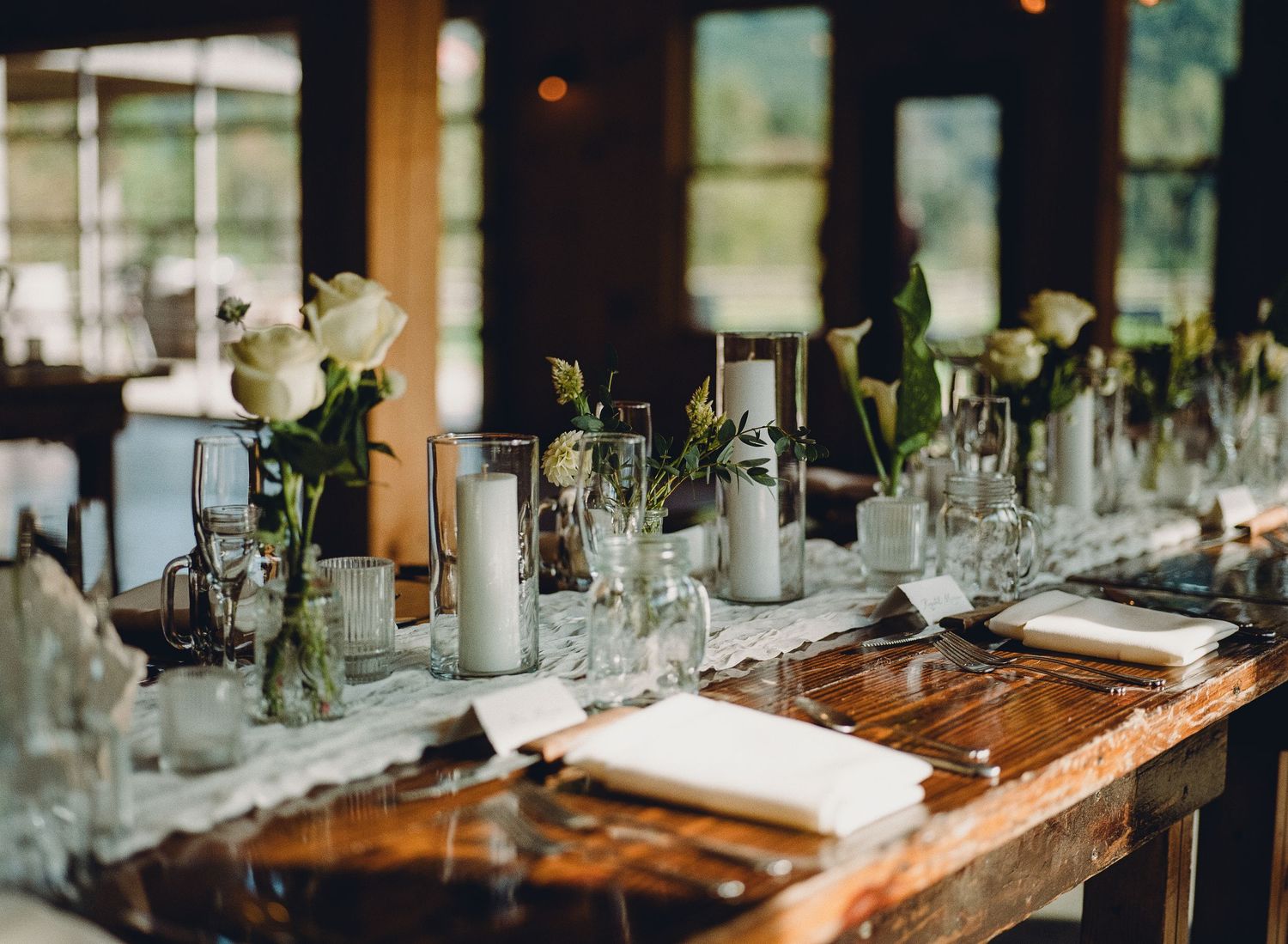 Elegant wedding reception table with white roses, silver candleholders, and place settings on a rustic wooden table.