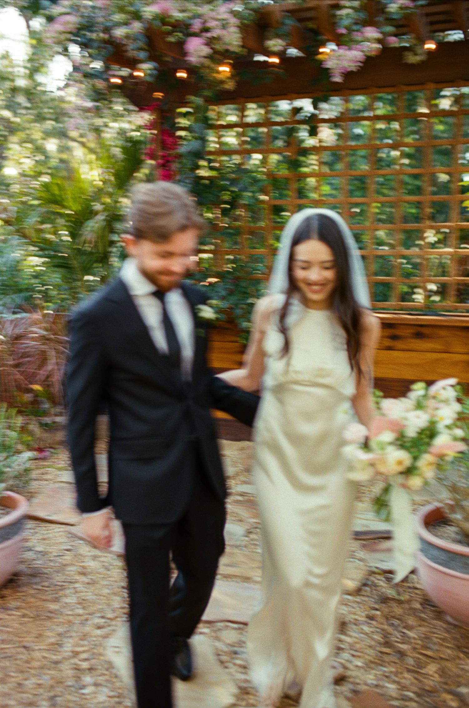A couple walks together through a garden pergola decorated with hanging flowers.